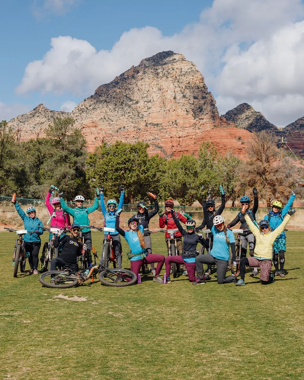 Group of women and men with mountain bikes, wearing helmets and outdoor gear, celebrating on a grassy field with rocky mountain and trees in the background.