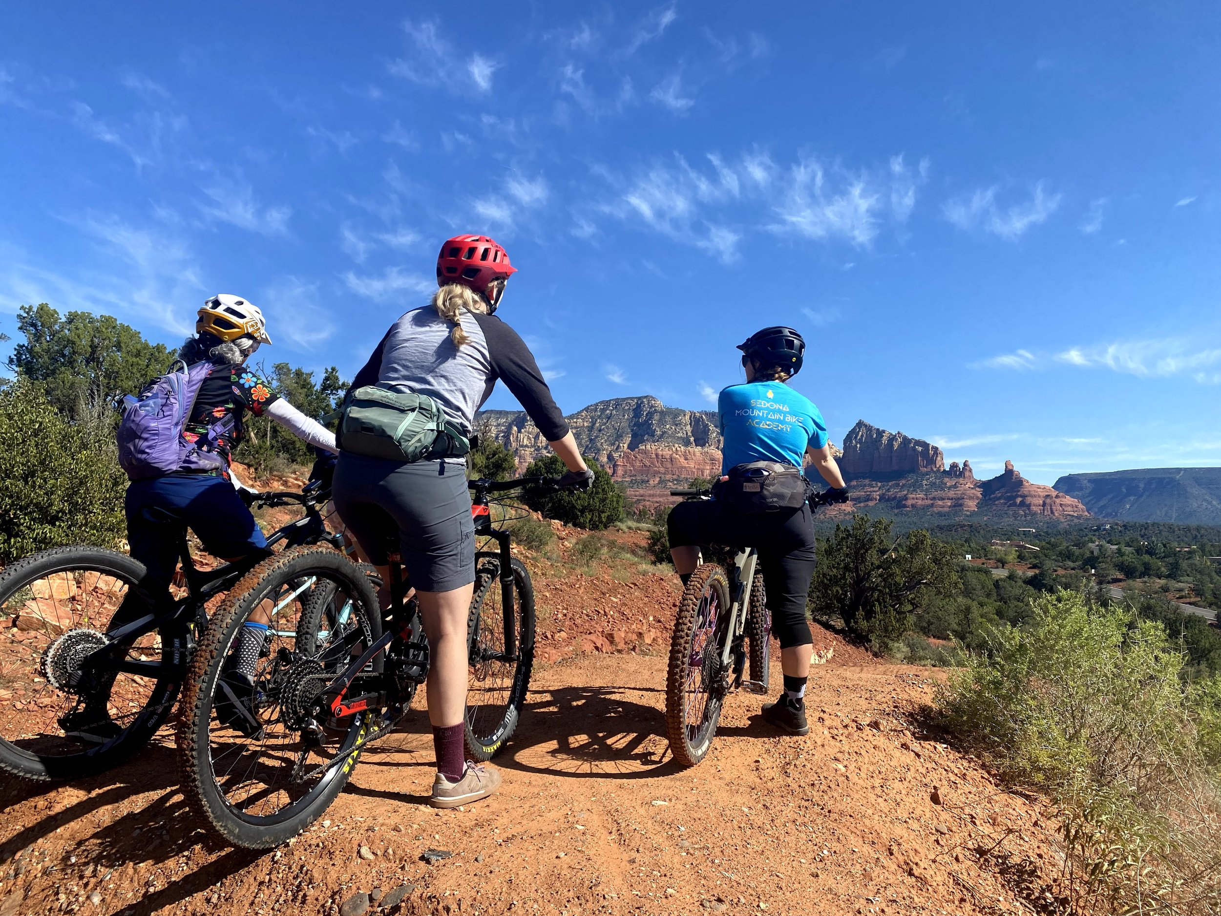 Three women with mountain bikes looking at scenery of red rock formations and distant mountains under a partly cloudy blue sky.