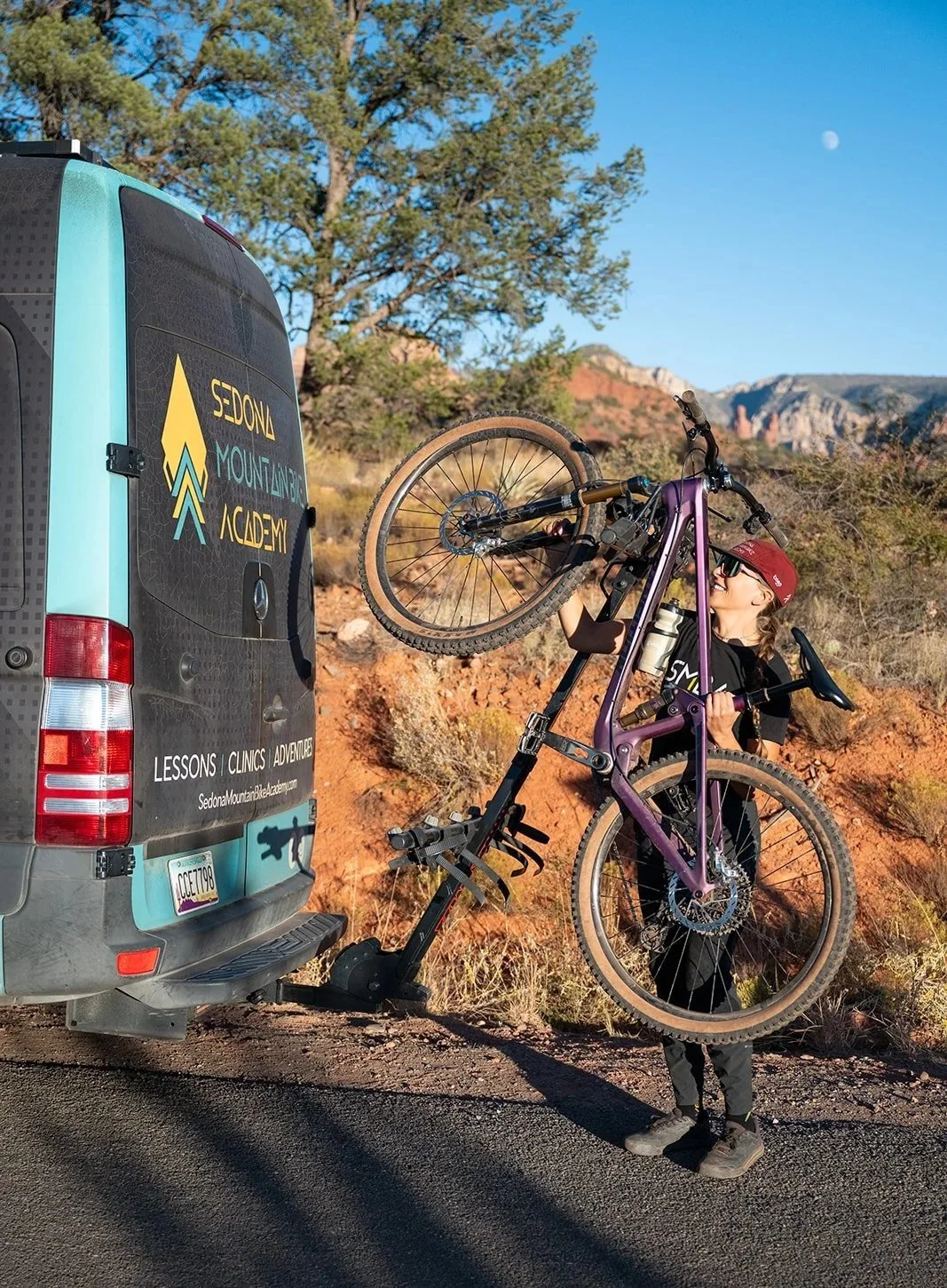 A woman loading a purple mountain bike onto a van with Sedona Mountain Bike Academy branding on the rear door, in a desert landscape with red rocks, shrubs, and a clear blue sky.