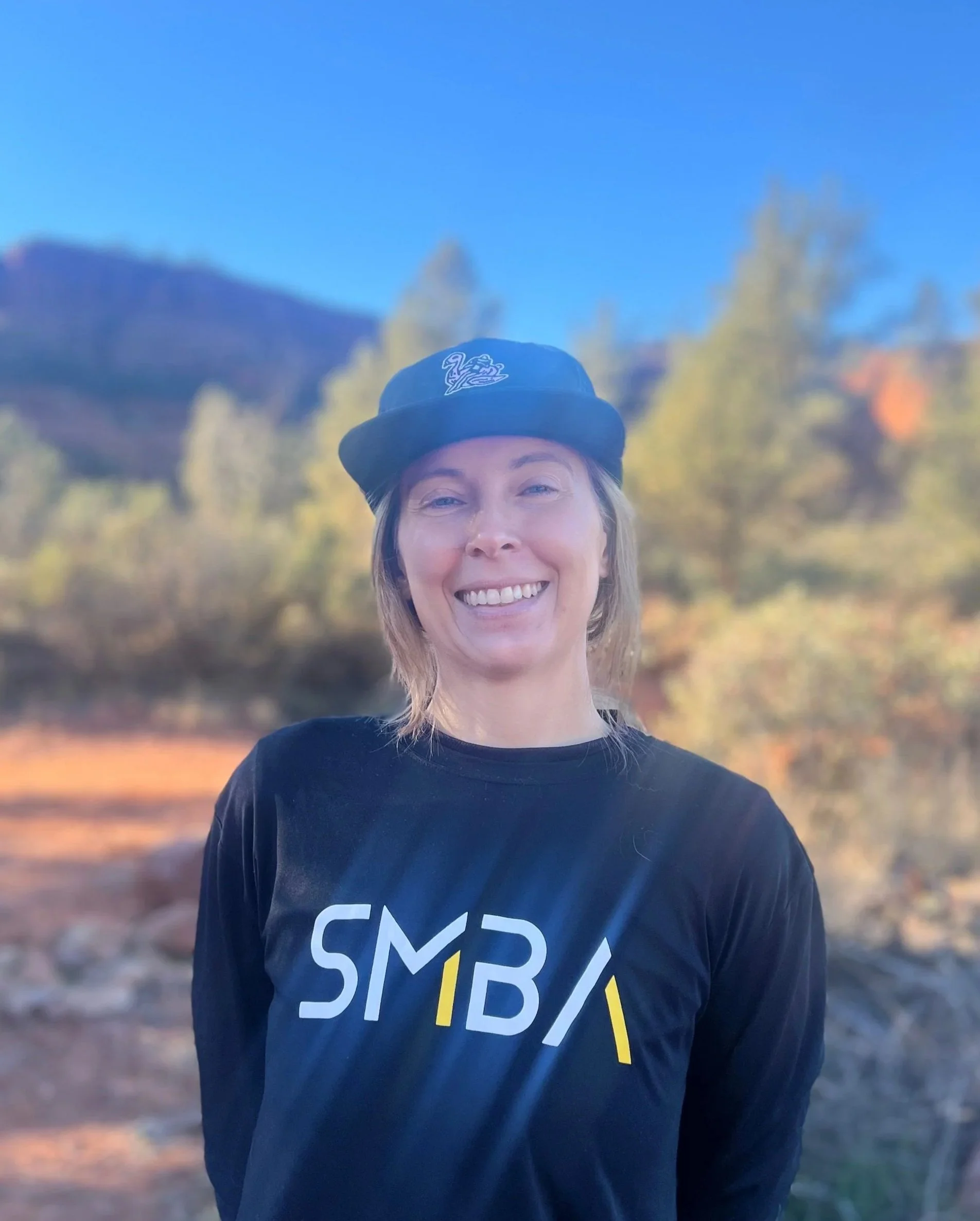 Smiling woman wearing a black cap with a white logo, standing outdoors with trees and mountain in the background, wearing a black T-shirt with white and yellow text.