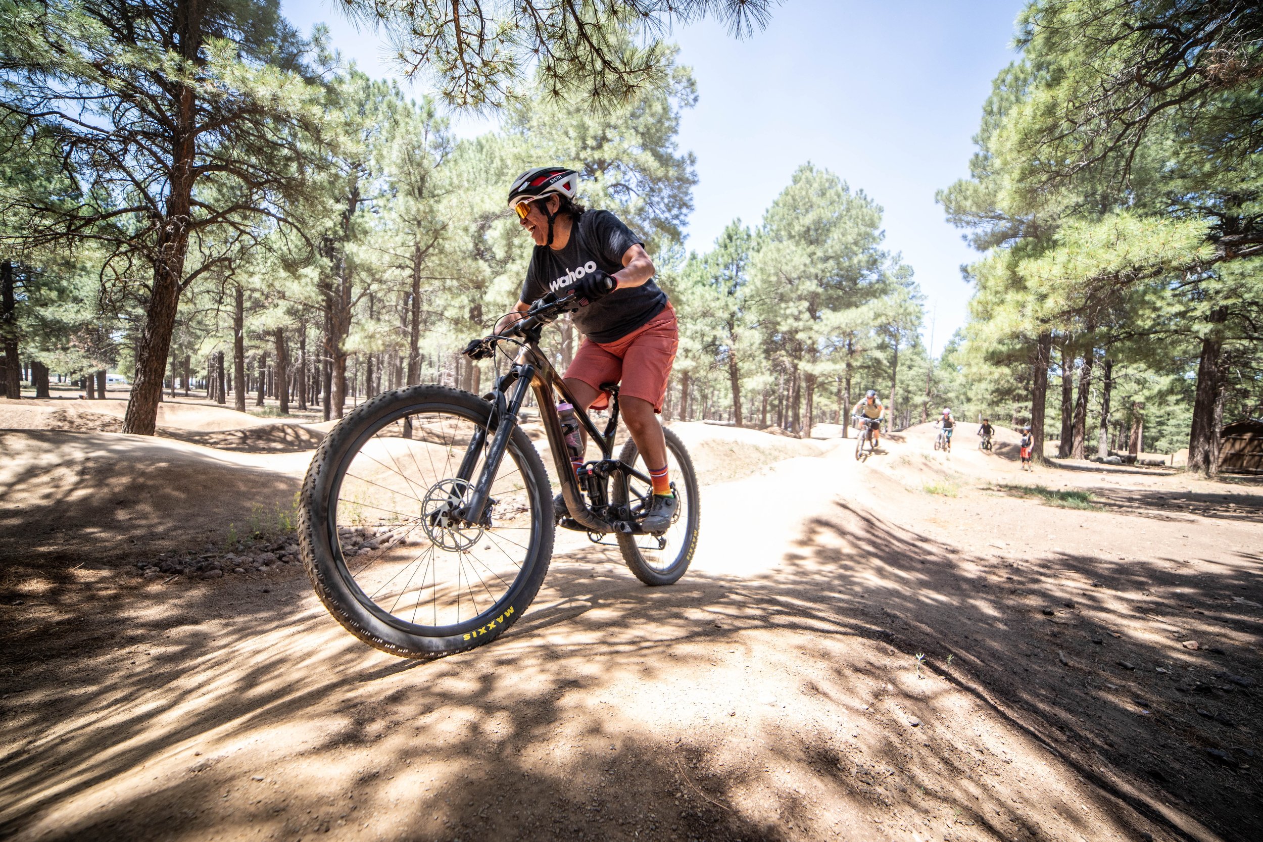 Person riding a mountain bike on a dirt trail in a forest with tall pine trees. The rider is wearing a helmet, sunglasses, a black T-shirt, and red shorts, smiling as they navigate the trail. Several other bikers are visible in the background.