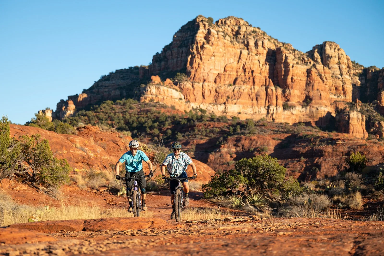 Two men riding mountain bikes on a trail in a desert landscape with red rock formations and sparse vegetation under a clear blue sky.
