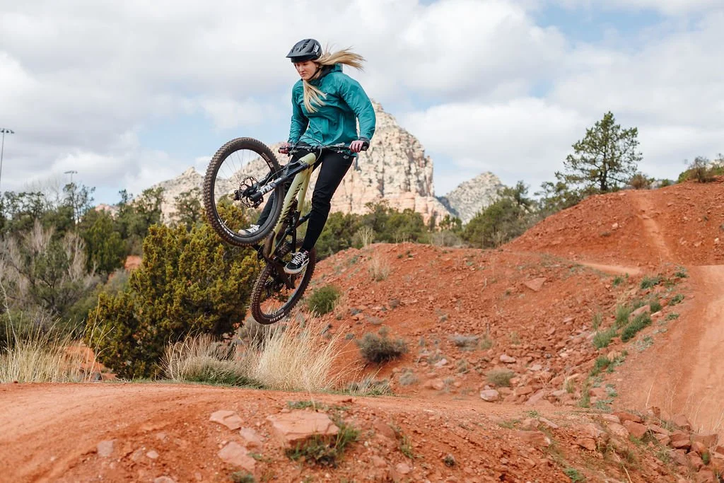 A woman in a blue jacket and black helmet jumping with her mountain bike on a dirt trail in a desert landscape with red rocks and sparse vegetation under an overcast sky.