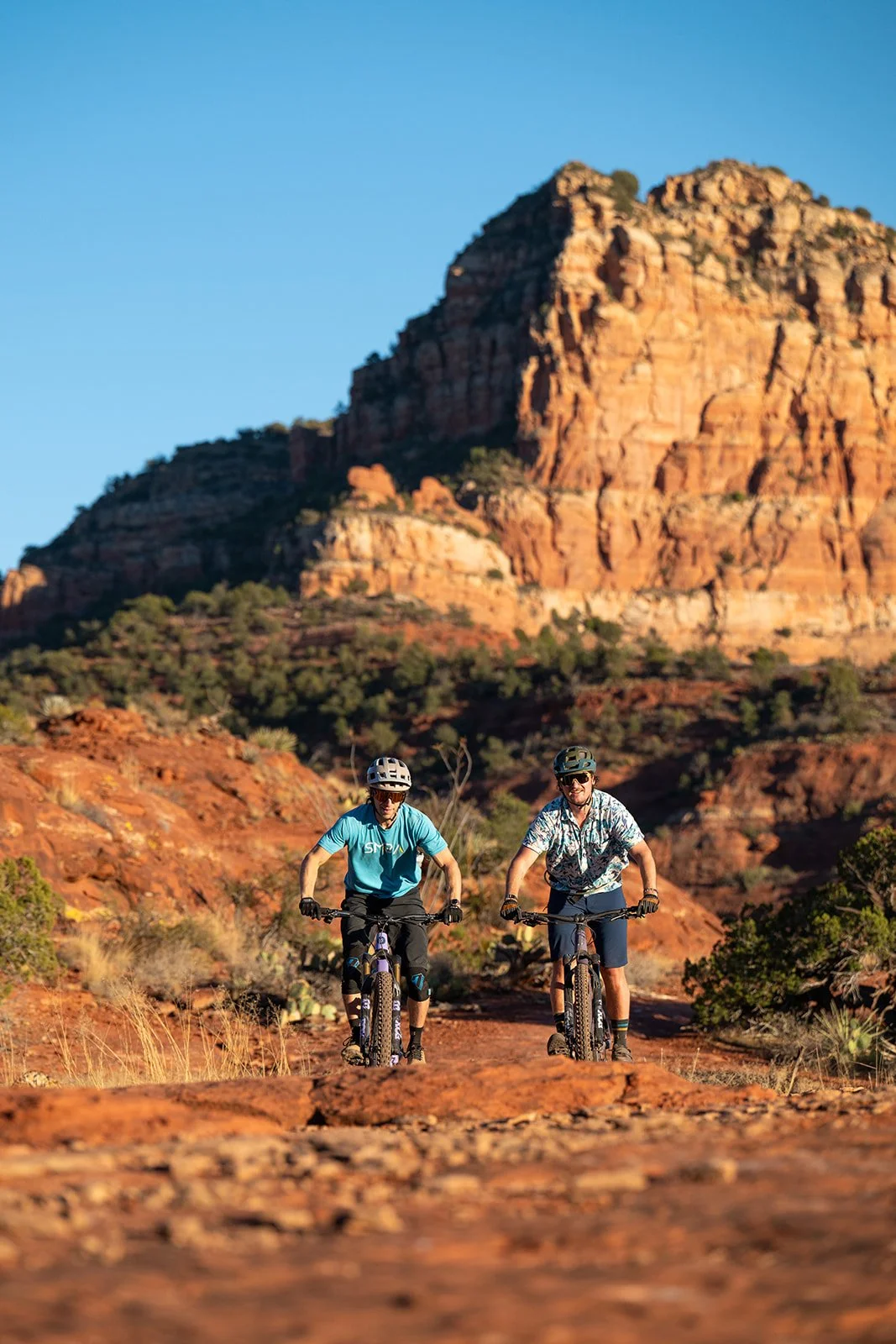 Two people riding mountain bikes on a dirt trail surrounded by red rocks and greenery in a desert landscape with a large rocky mountain in the background.
