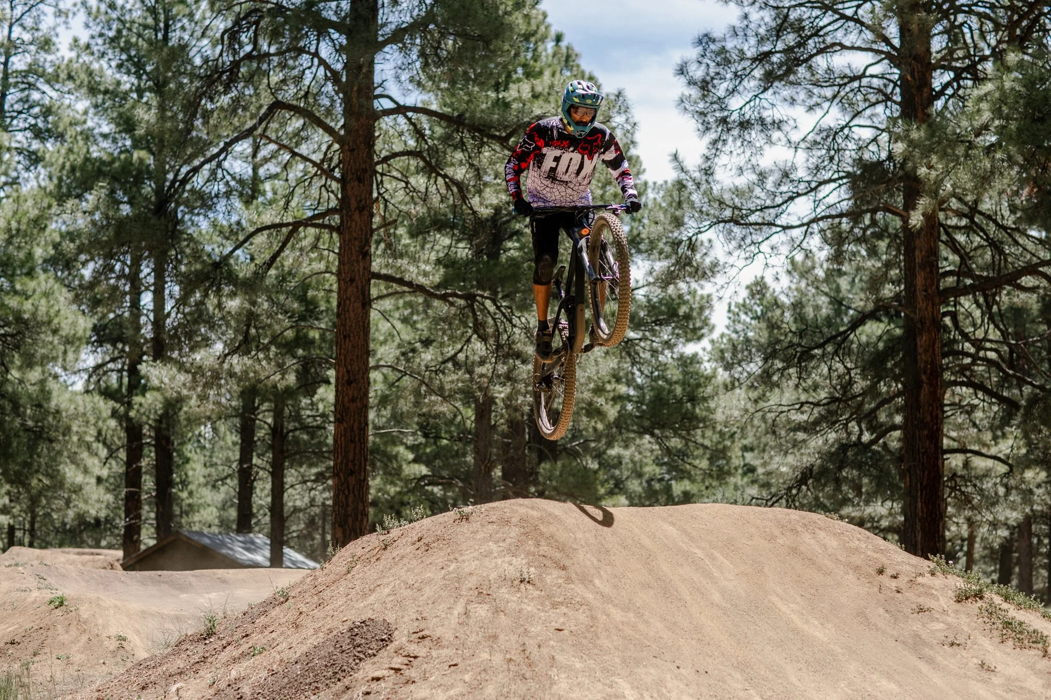 A mountain biker wearing a helmet and jersey is airborne above a dirt ramp in a forested area.