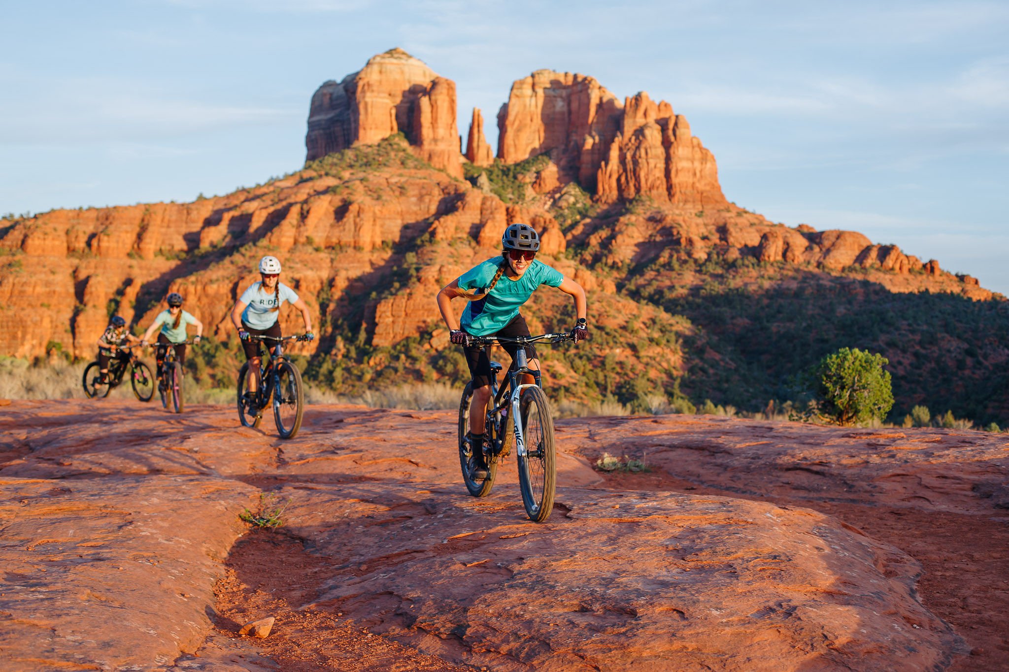 Four people riding mountain bikes in Sedona on a red rocky trail with red rock formations in the background.