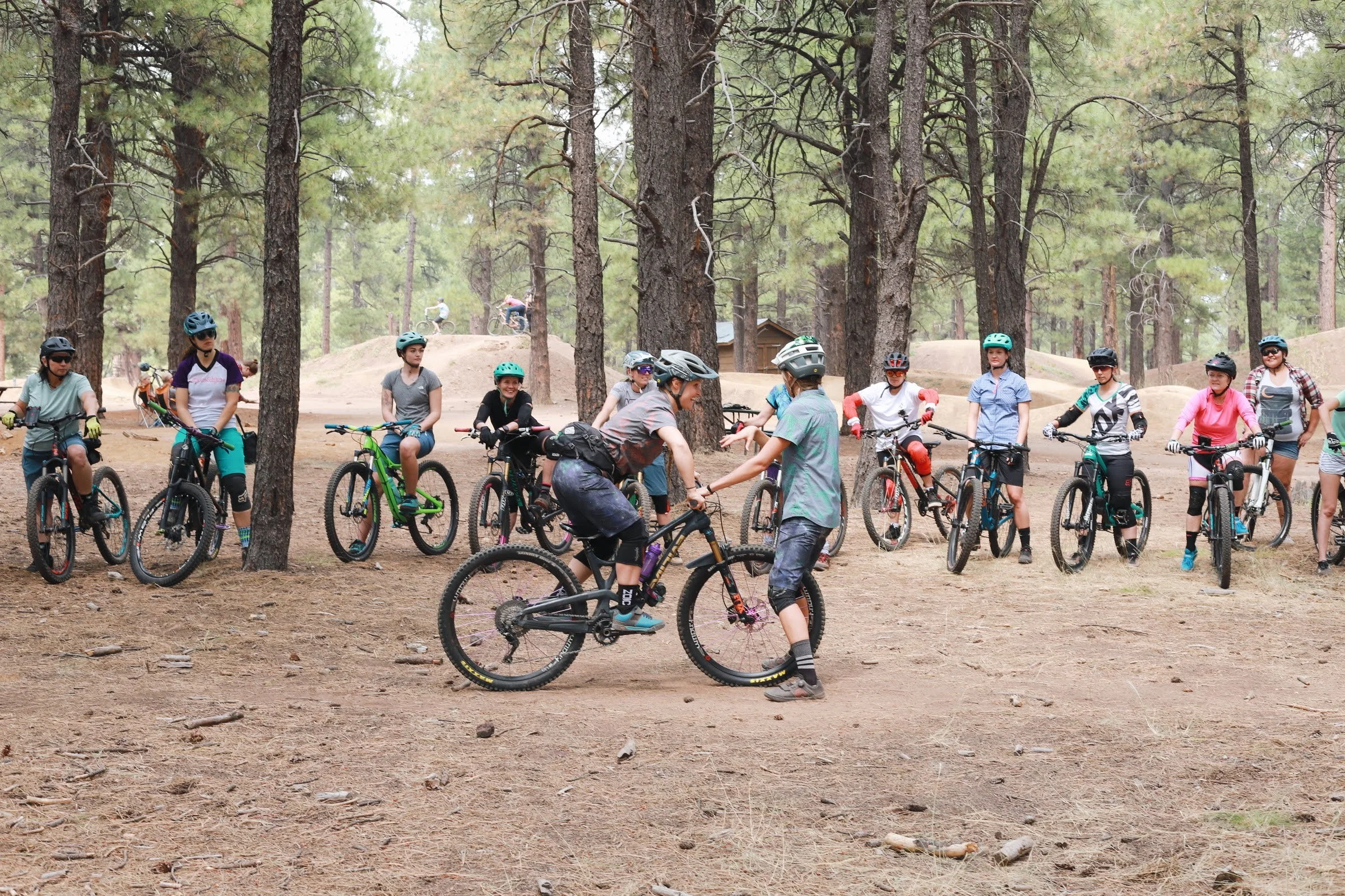 A group of women mountain bikers gathered in a forest, wearing helmets, learning from two mountain bike instructors, amongst tall trees.