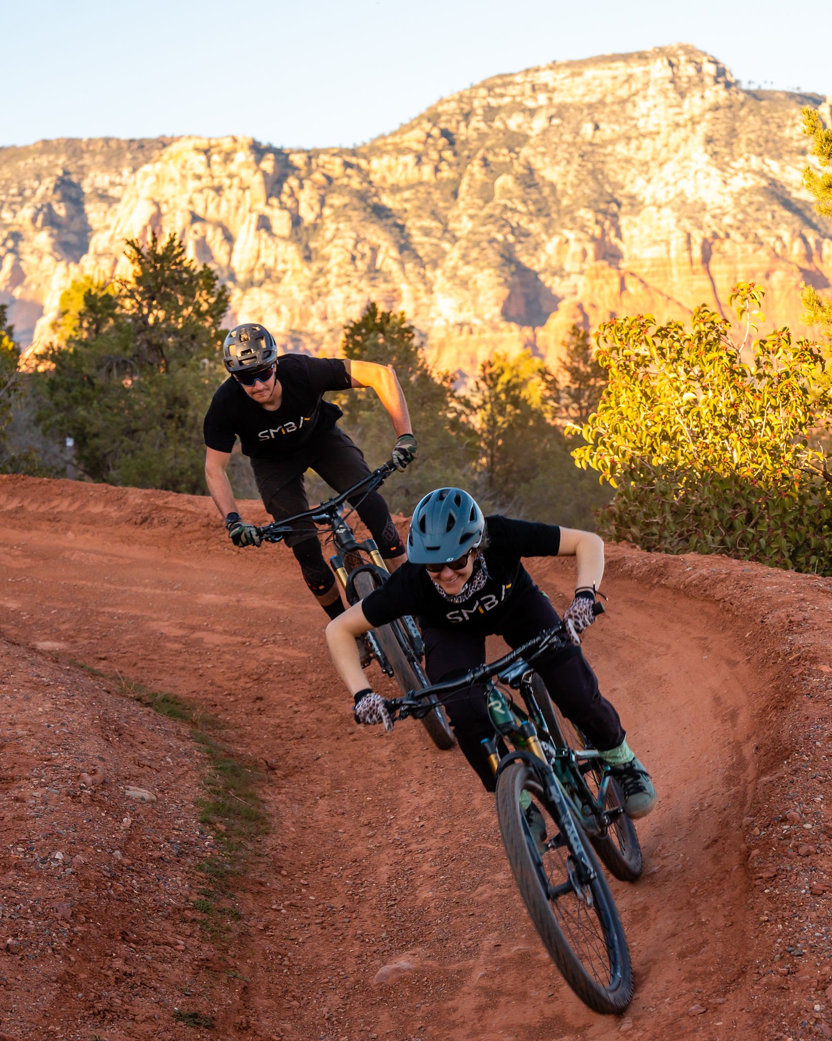 Two mountain bikers riding on a dirt trail in a desert landscape with large rock formations and trees, during sunset.