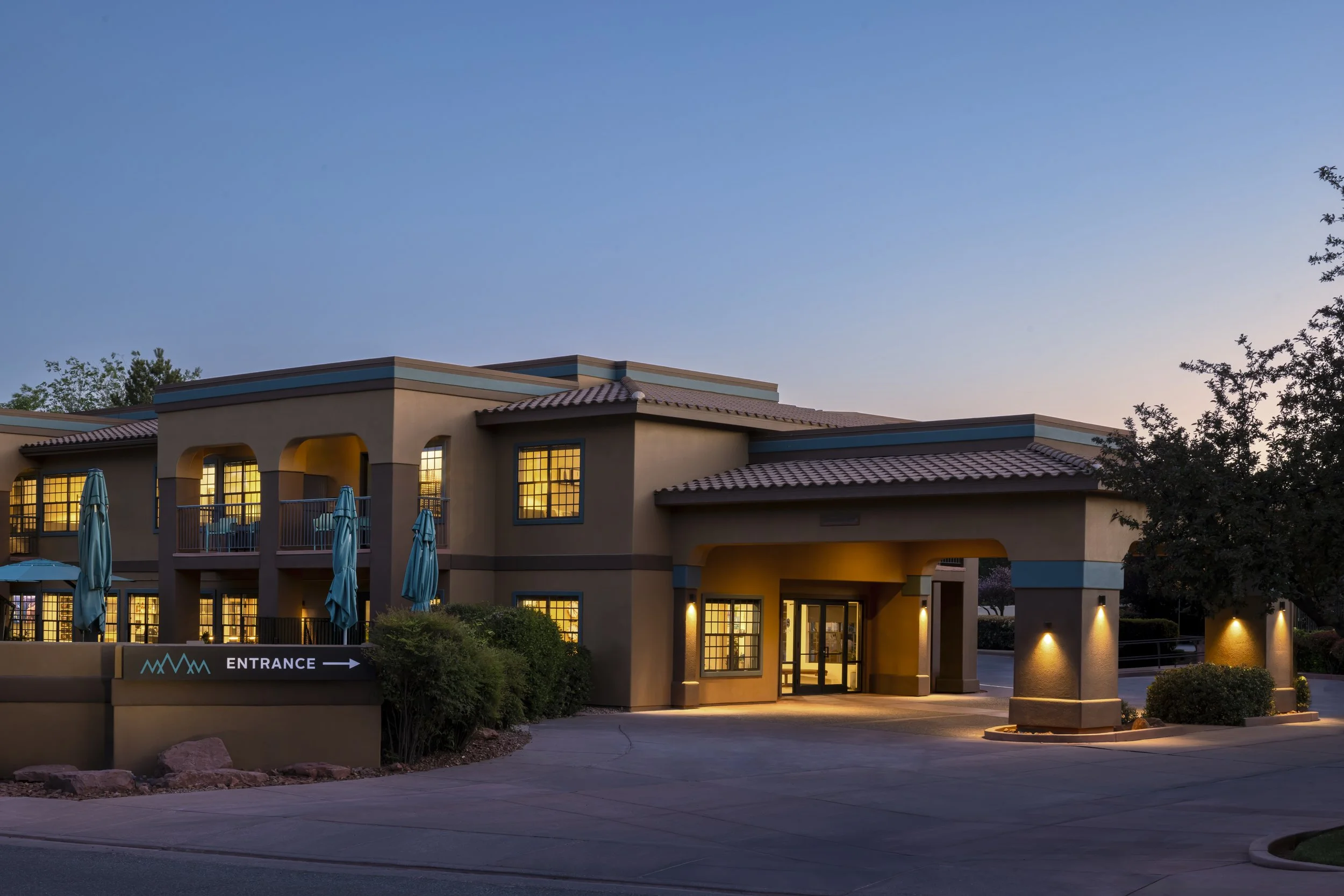 A modern two-story hotel is Sedona, Arizona with balconies, large windows, and a tiled roof, illuminated by exterior lighting during sunset, with a sign indicating the entrance.