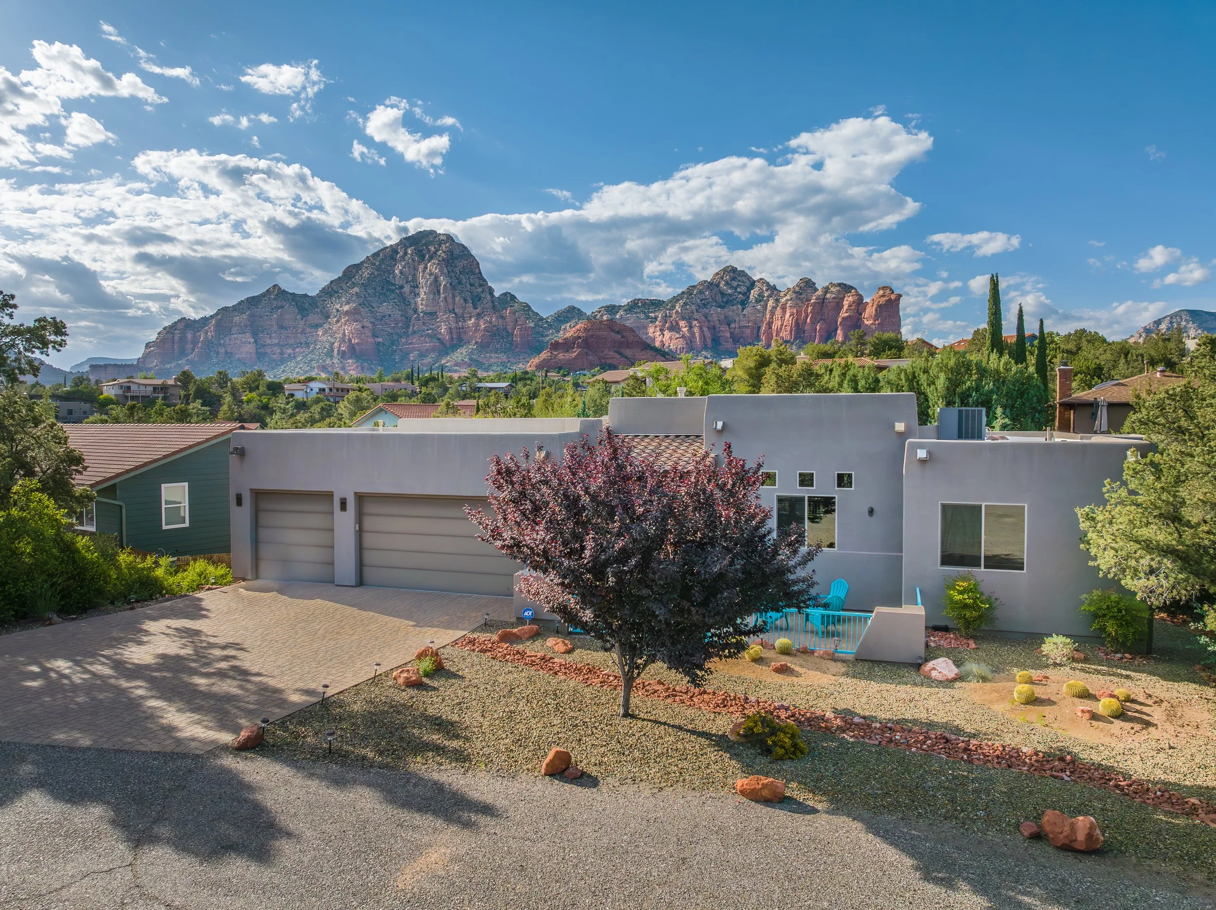 Residential house with a front yard, a purple-leafed tree, and desert landscaping, set against a backdrop of red-rock mountains under a partly cloudy sky.