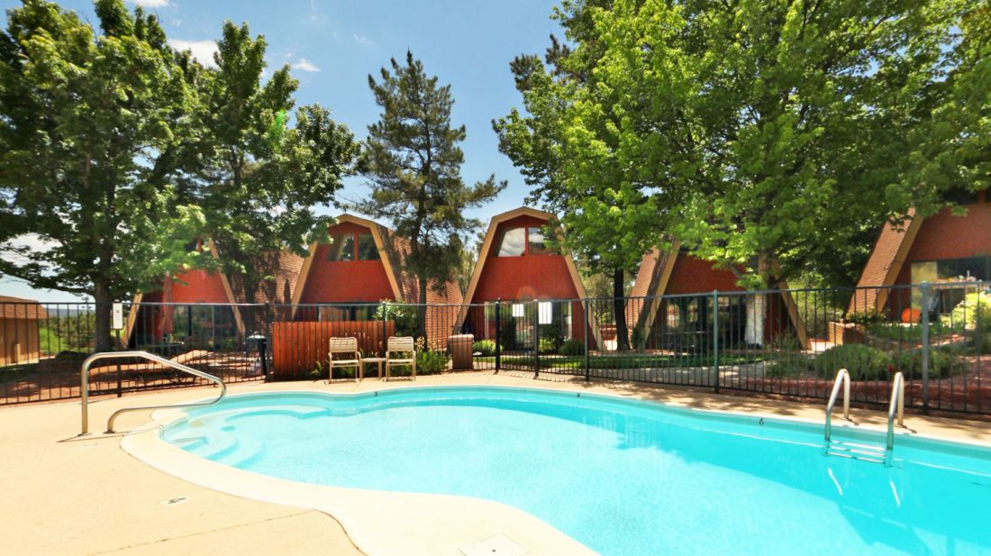 An outdoor swimming pool surrounded by a black metal fence, with several Adirondack chairs nearby, tall green trees, and brown A-frame style houses in the background, under a partly cloudy blue sky.
