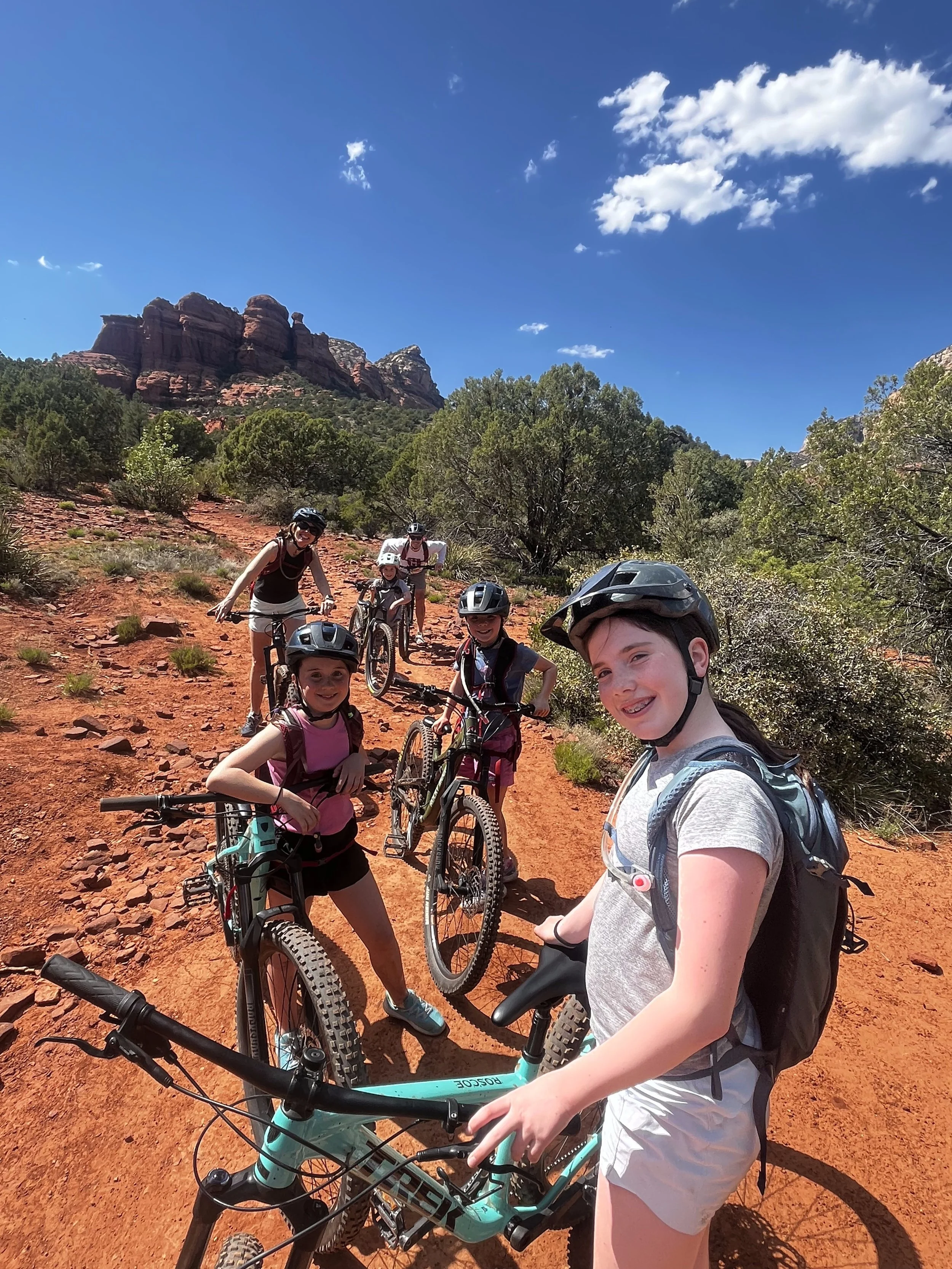Group of children and adults mountain biking in Sedona on a dirt trail through a desert landscape with red rock formations, green bushes, and a blue sky with clouds.