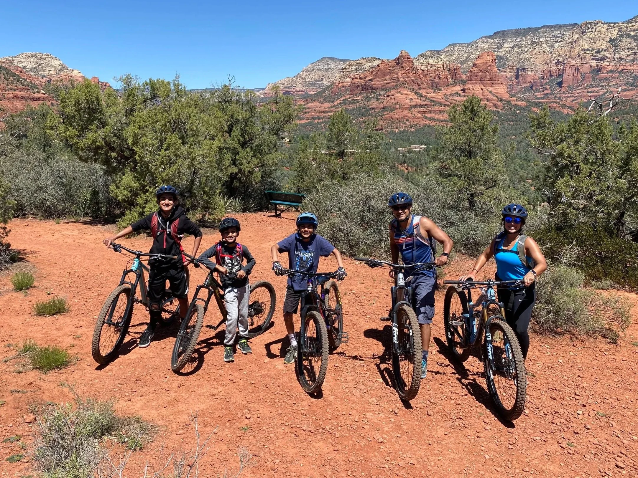 Group of five people in cycling gear with mountain bikes on a dirt trail in a desert landscape with red rock formations and green shrubbery in the background.
