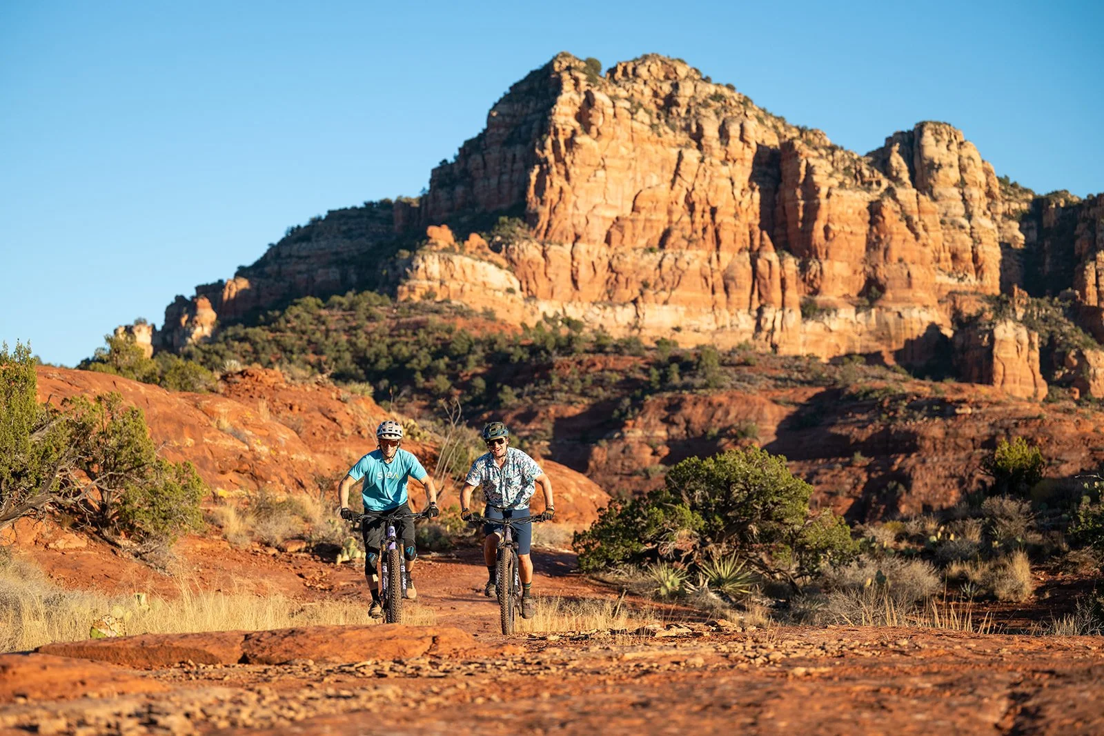 Two men riding mountain bikes on a desert trail with red rocks and a large rock formation in the background.