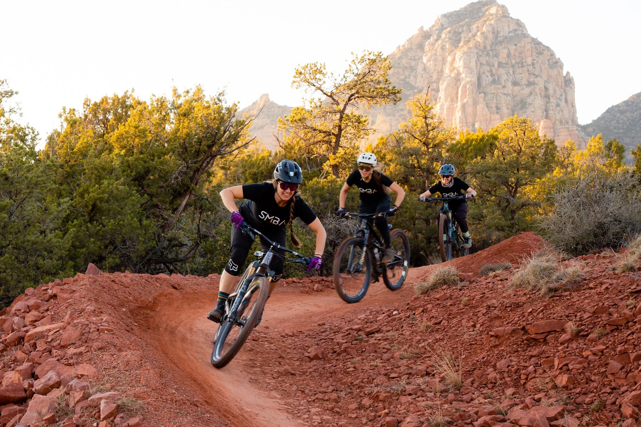 Three women mountain biking on a dirt trail surrounded by trees and red rocks, with a large mountain in the background during sunset.