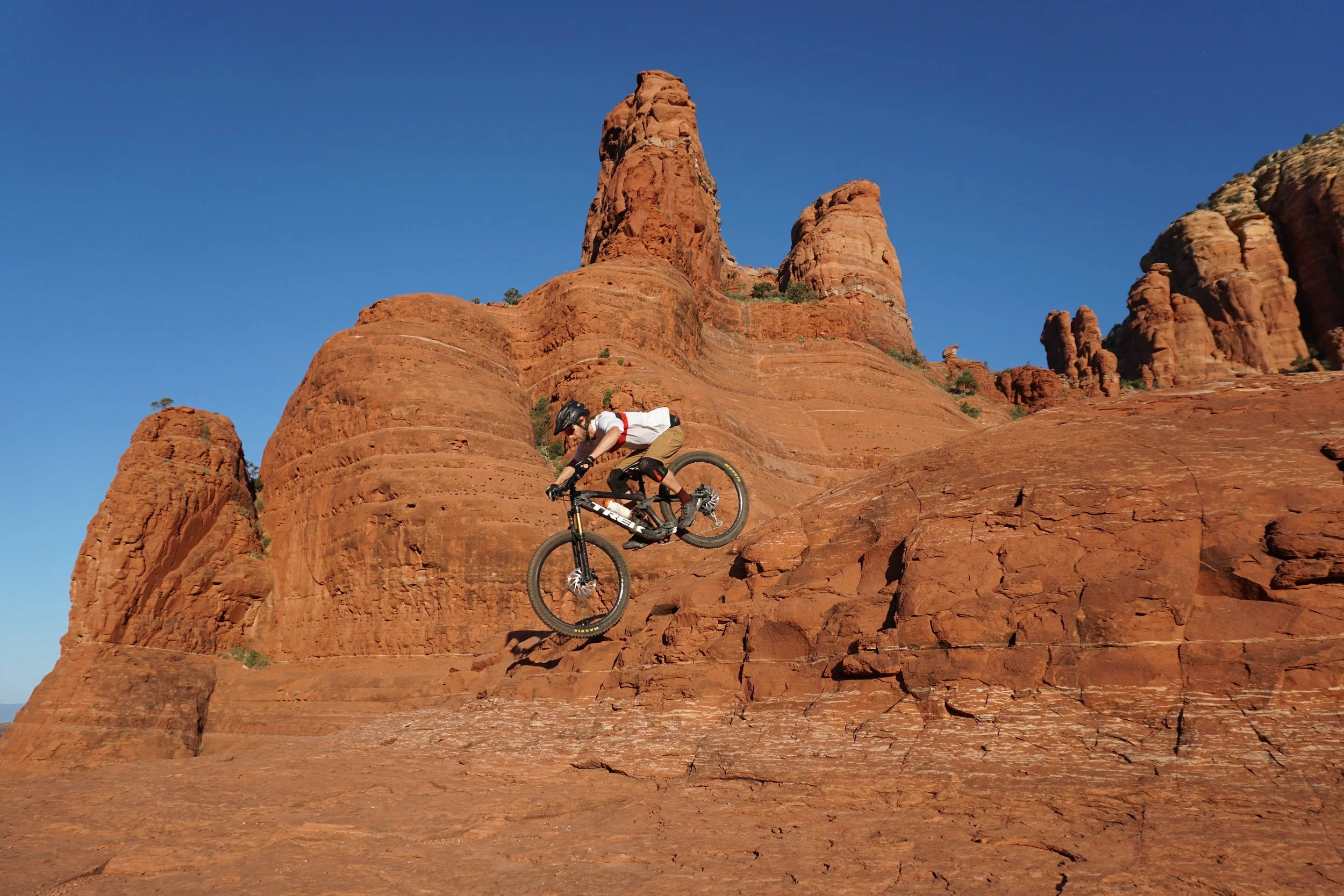 A person riding a mountain bike down a red rock slope in a desert landscape with tall red rock formations and a clear blue sky.