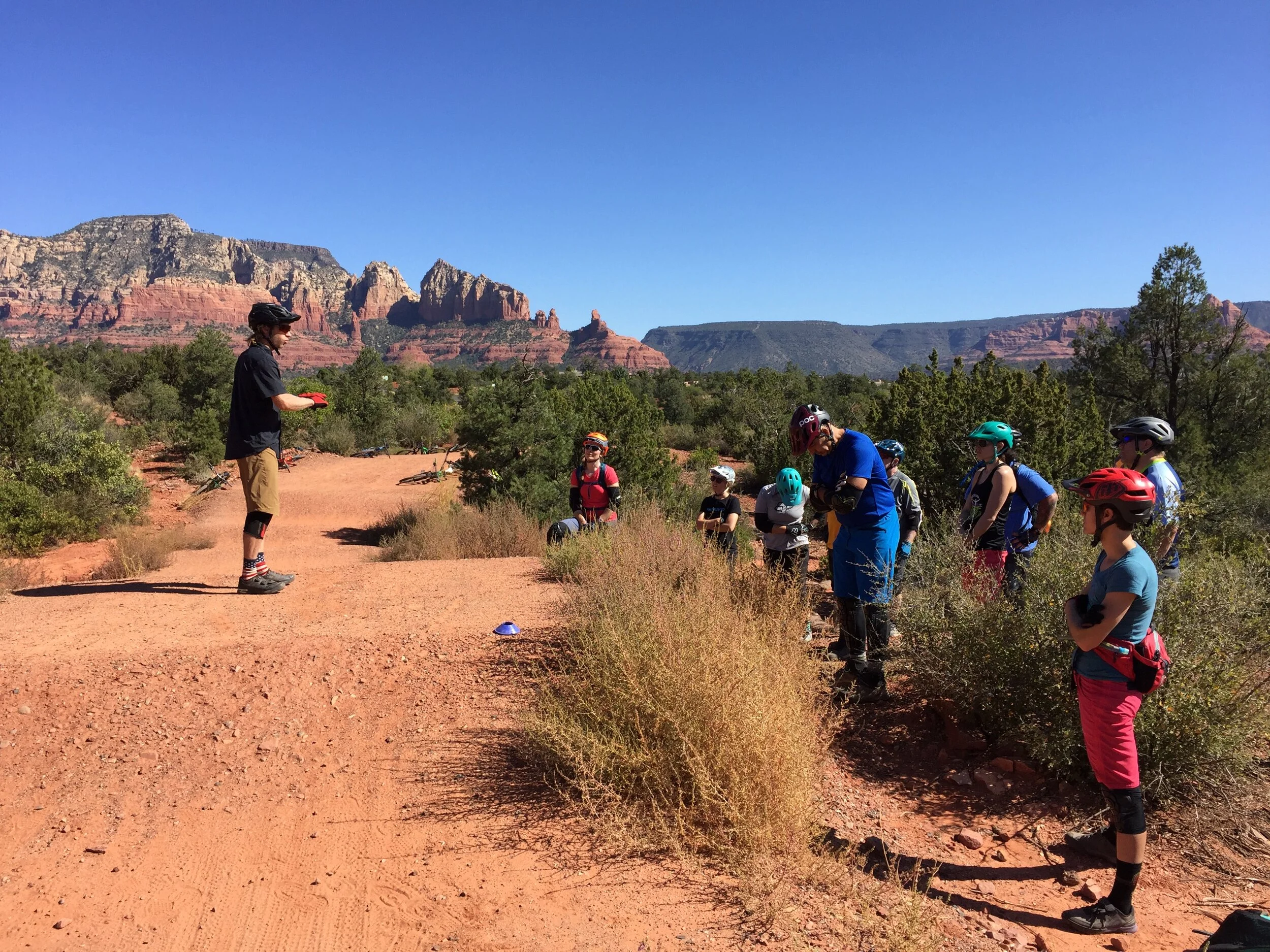 Group of mountain bikers in outdoor gear, with helmets and knee pads, standing on a dirt trail in a desert landscape with red rock formations and sparse vegetation under a clear blue sky, participating in a mountain bike clinic.