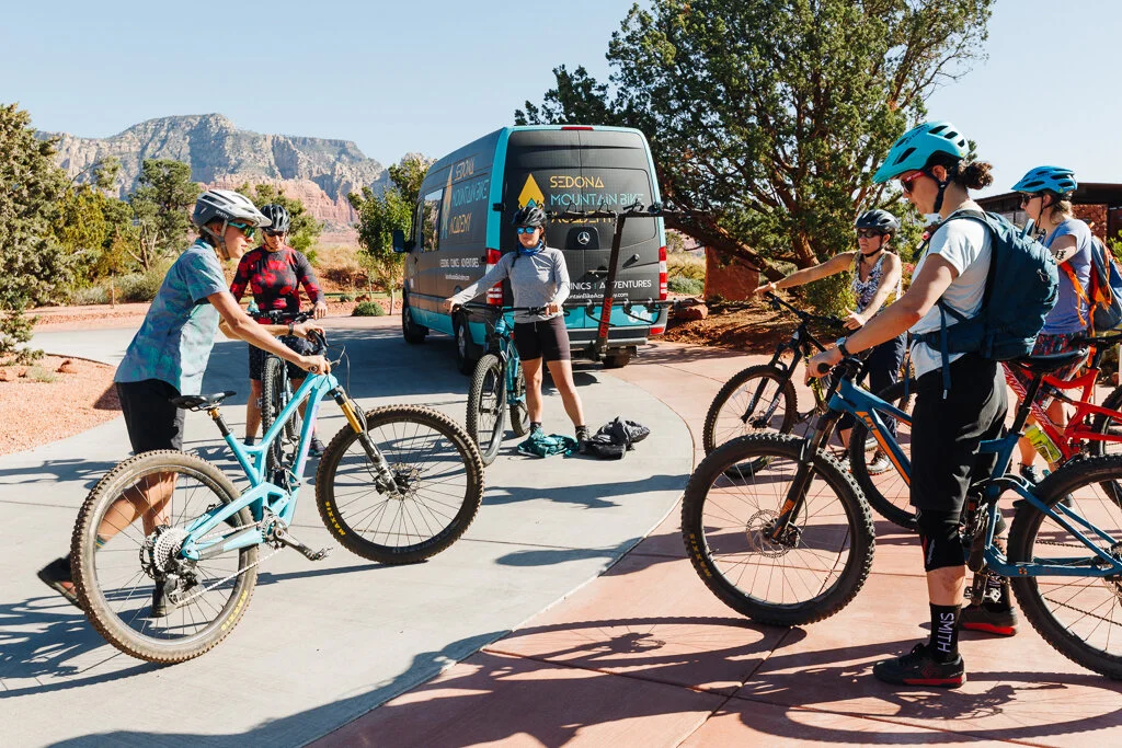 Group of mountain bikers wearing helmets, gathered around with bikes, preparing for a ride at a scenic trailhead with a van and red rock formations in the background.