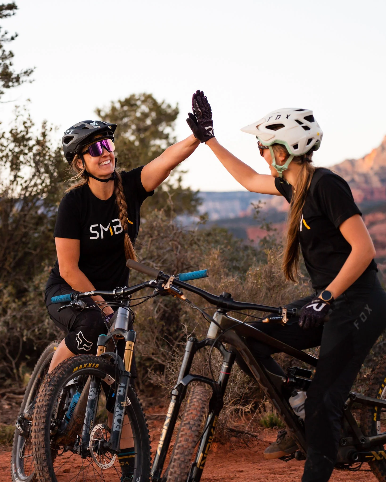 Two women wearing mountain biking helmets and gloves high-five while riding bikes on a dirt trail with trees and mountains in the background.