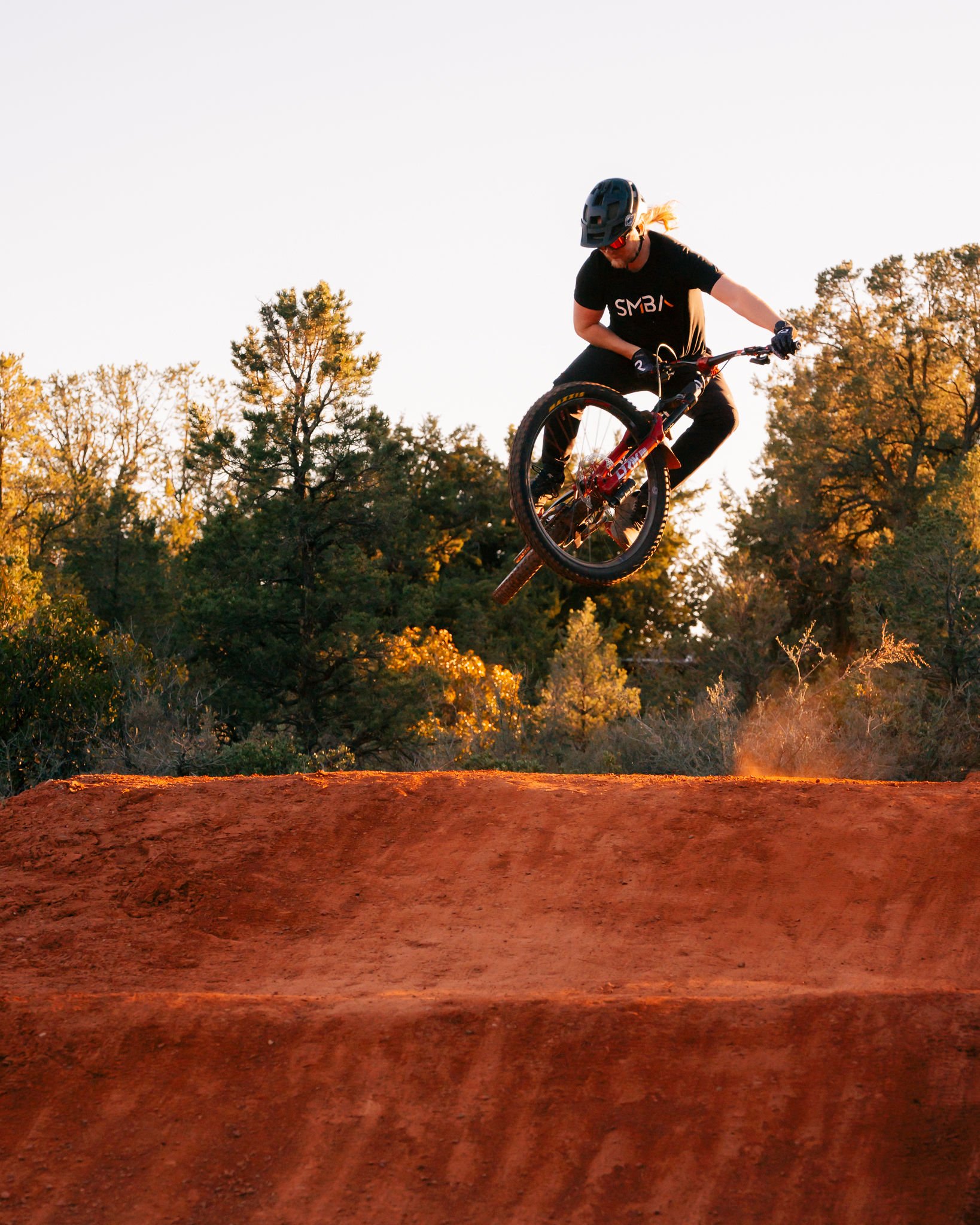 A person wearing a black helmet and black shirt riding a mountain bike on a dirt trail during sunset, with trees and clear sky in the background.
