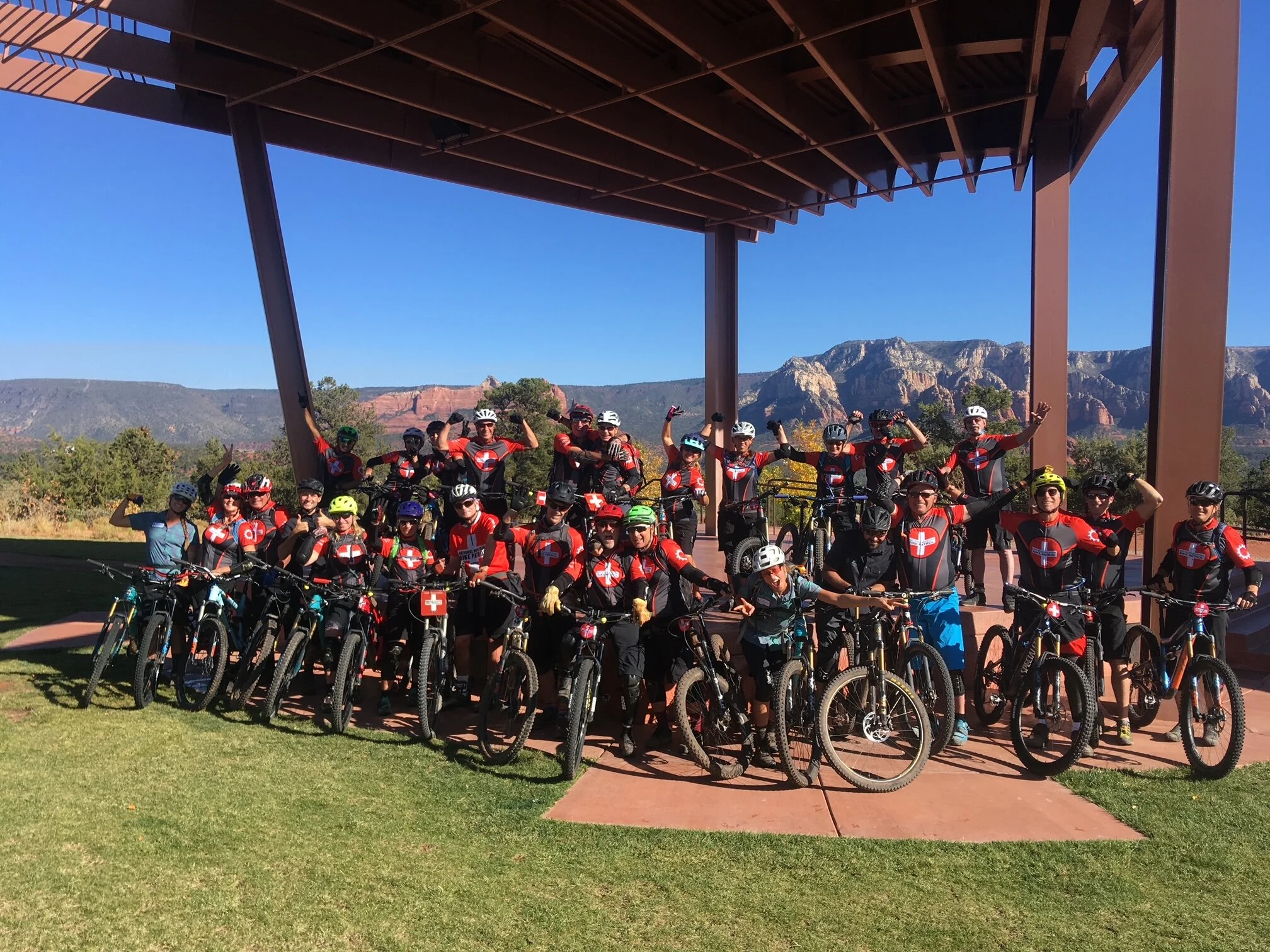 Group of cyclists in matching red and black jerseys, some raising their arms, posing with mountain bikes under a large metal pavilion against a backdrop of mountains and clear blue sky.