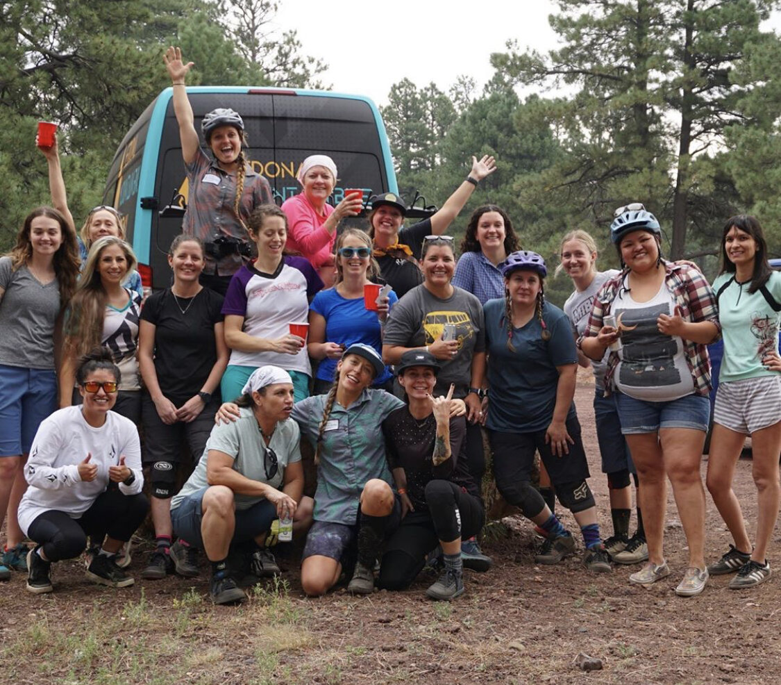 A group of female mountain bikers outdoors in front of a blue van surrounded by trees. Some are wearing helmets, and they are smiling, holding cups, and posing for the photo.