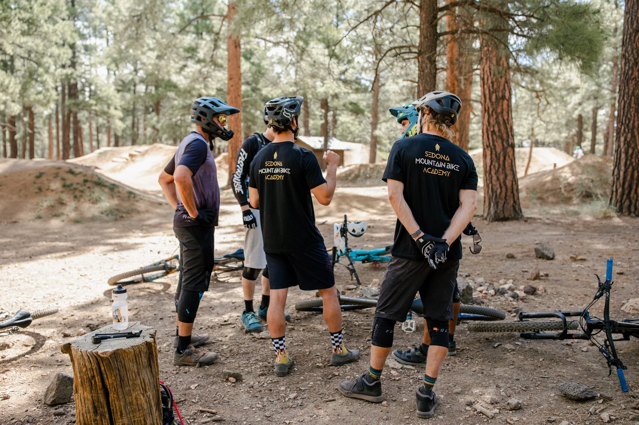 Group of five mountain bikers wearing helmets and mountain biking gear, standing together in a forested area with dirt trails and trees, preparing for or taking a break from riding.