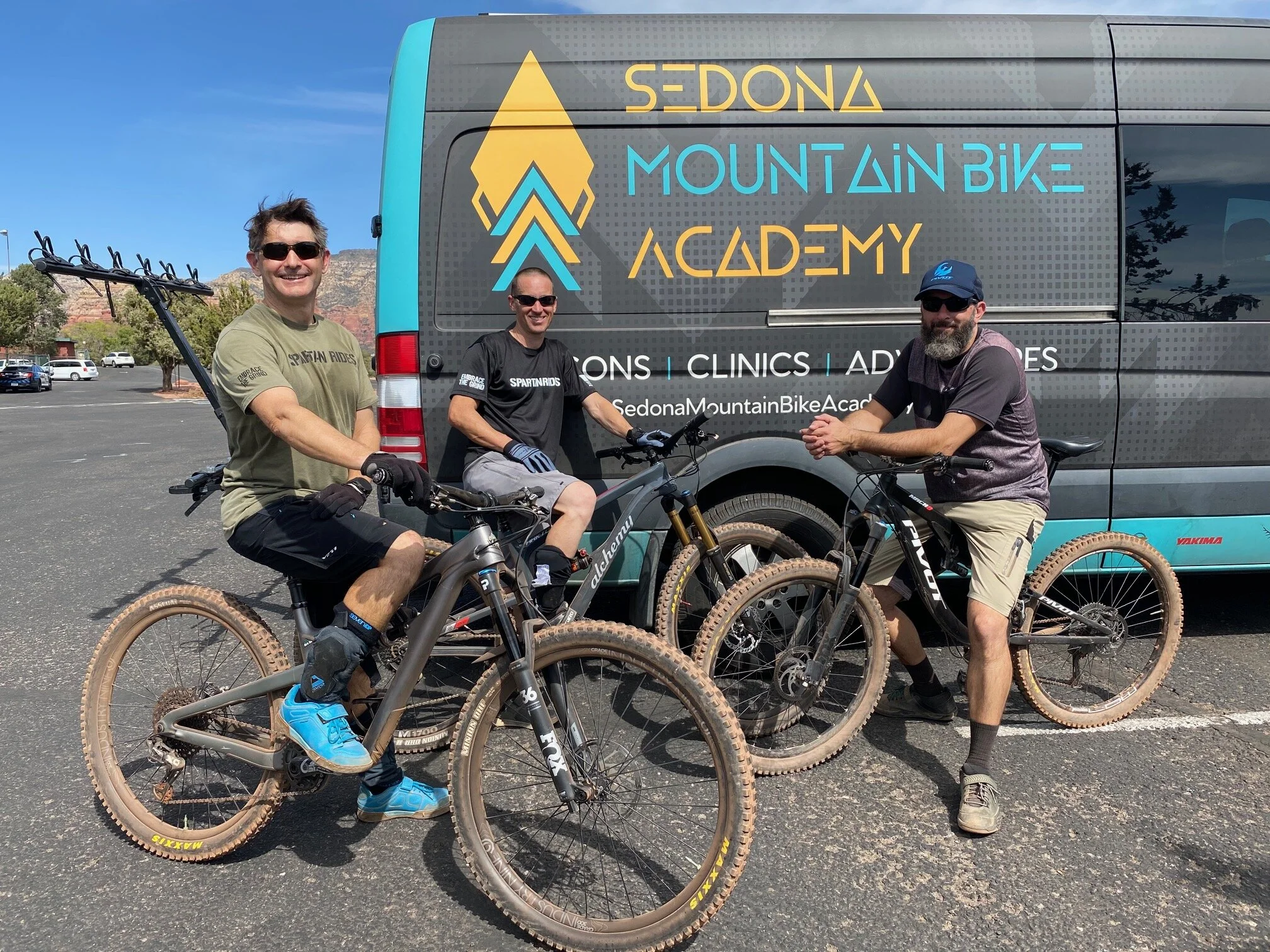 Three men with bicycles standing in front of a Sprinter van with the logos and text 'Sedona Mountain Bike Academy'  on it, in a parking lot under a clear sky.
