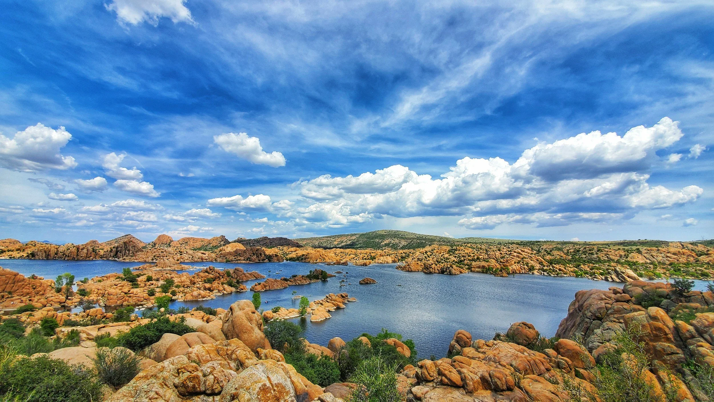 A landscape with rocky terrain, shrubs, and trees in the foreground, a body of water, and hills in the background under a partly cloudy blue sky.