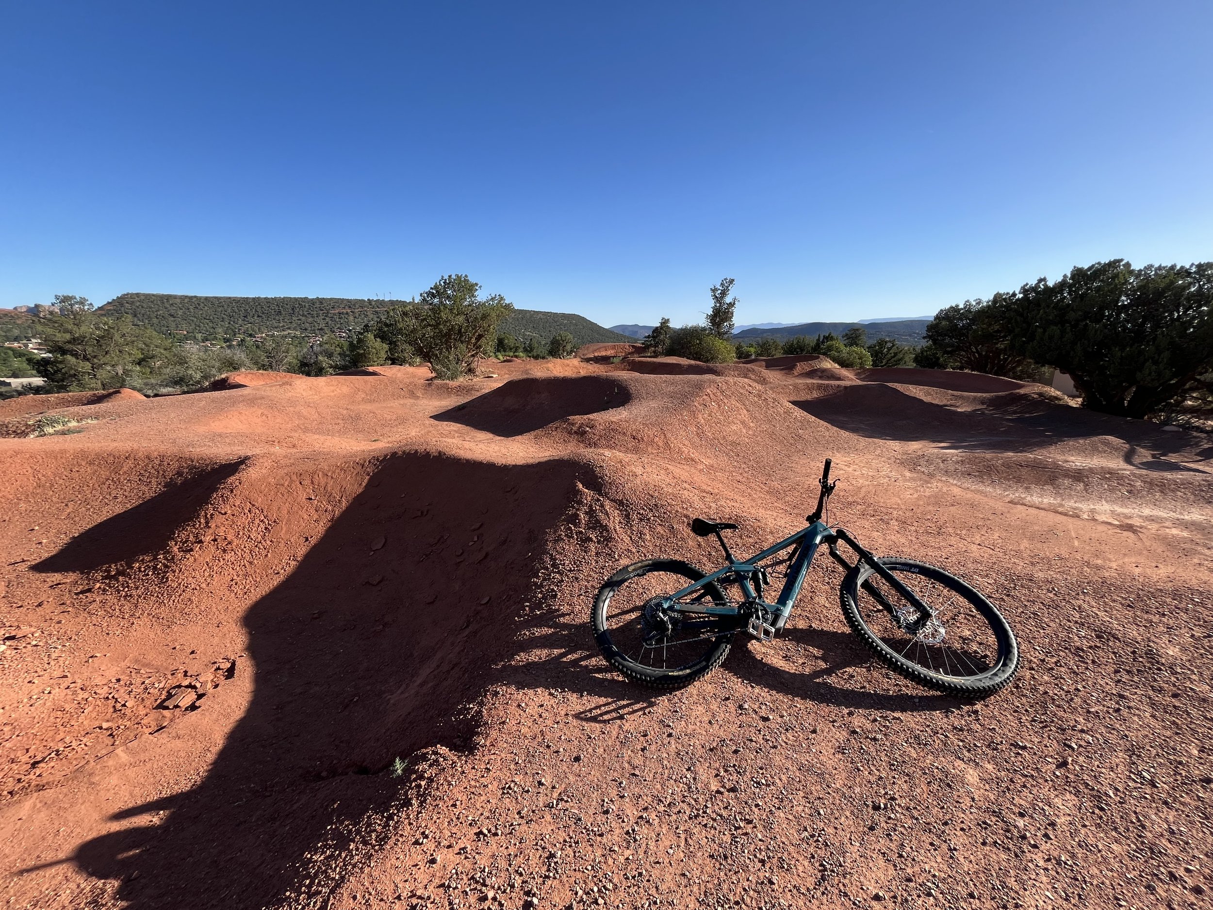 An e-mountain bike lying on its side on red dirt ground with hilly landscape and clear blue sky in the background.