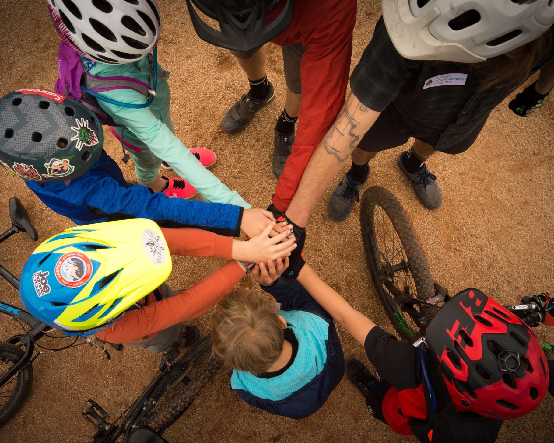 A group of children and adults in helmets and outdoor clothing gathered in a circle on a dirt surface, placing their hands together in a show of team spirit during a mountain biking activity.