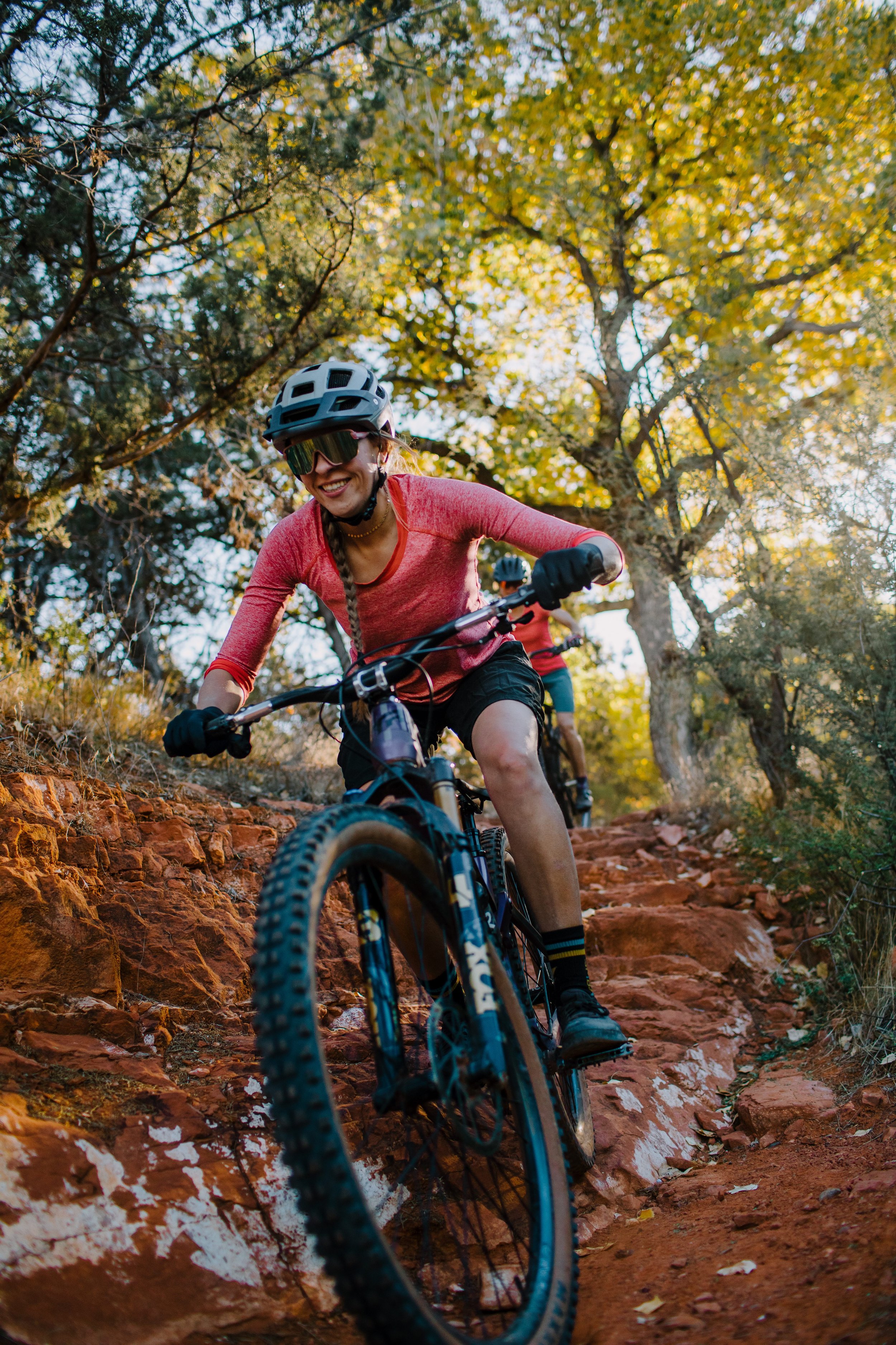 A woman mountain biking on a rocky trail through a forest with autumn-colored leaves, wearing a helmet and sunglasses, smiling.
