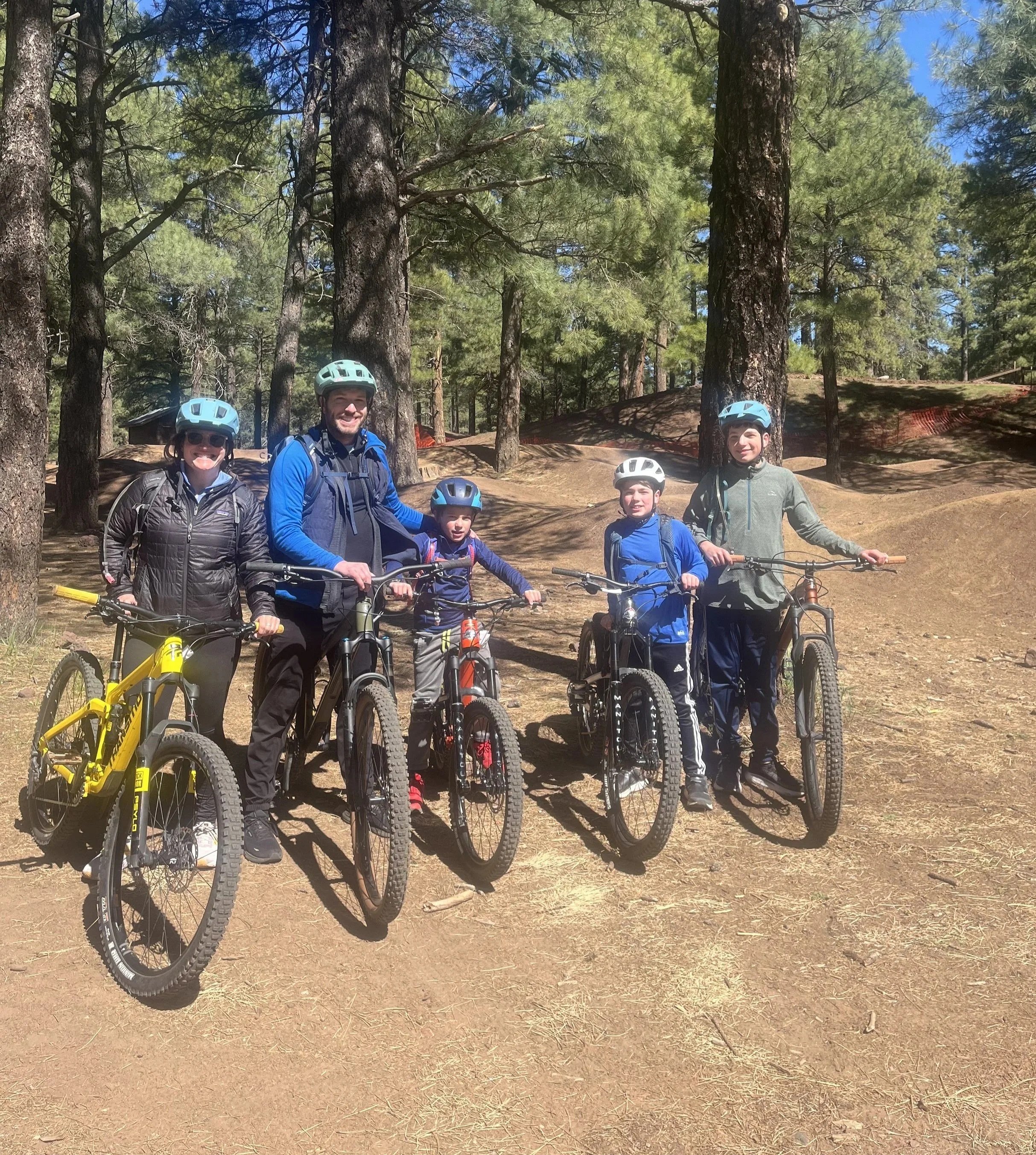 A family of five, wearing helmets and outdoor clothing, standing with their bicycles in a forested area with tall trees and a dirt ground.