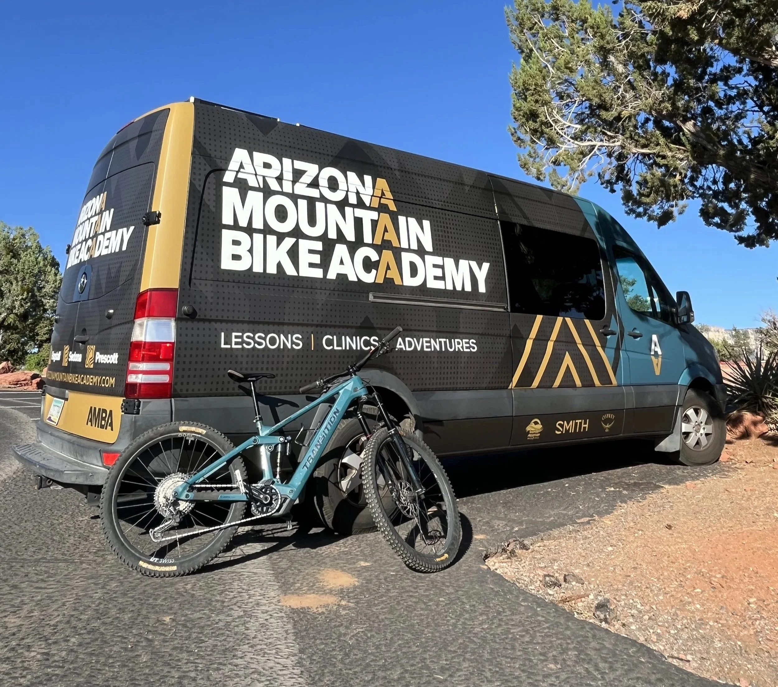 A black van with markings of Arizona Mountain Bike Academy parked outdoors with a blue sky and trees in the background. There is a teal e-mountain bike leaning against the van.
