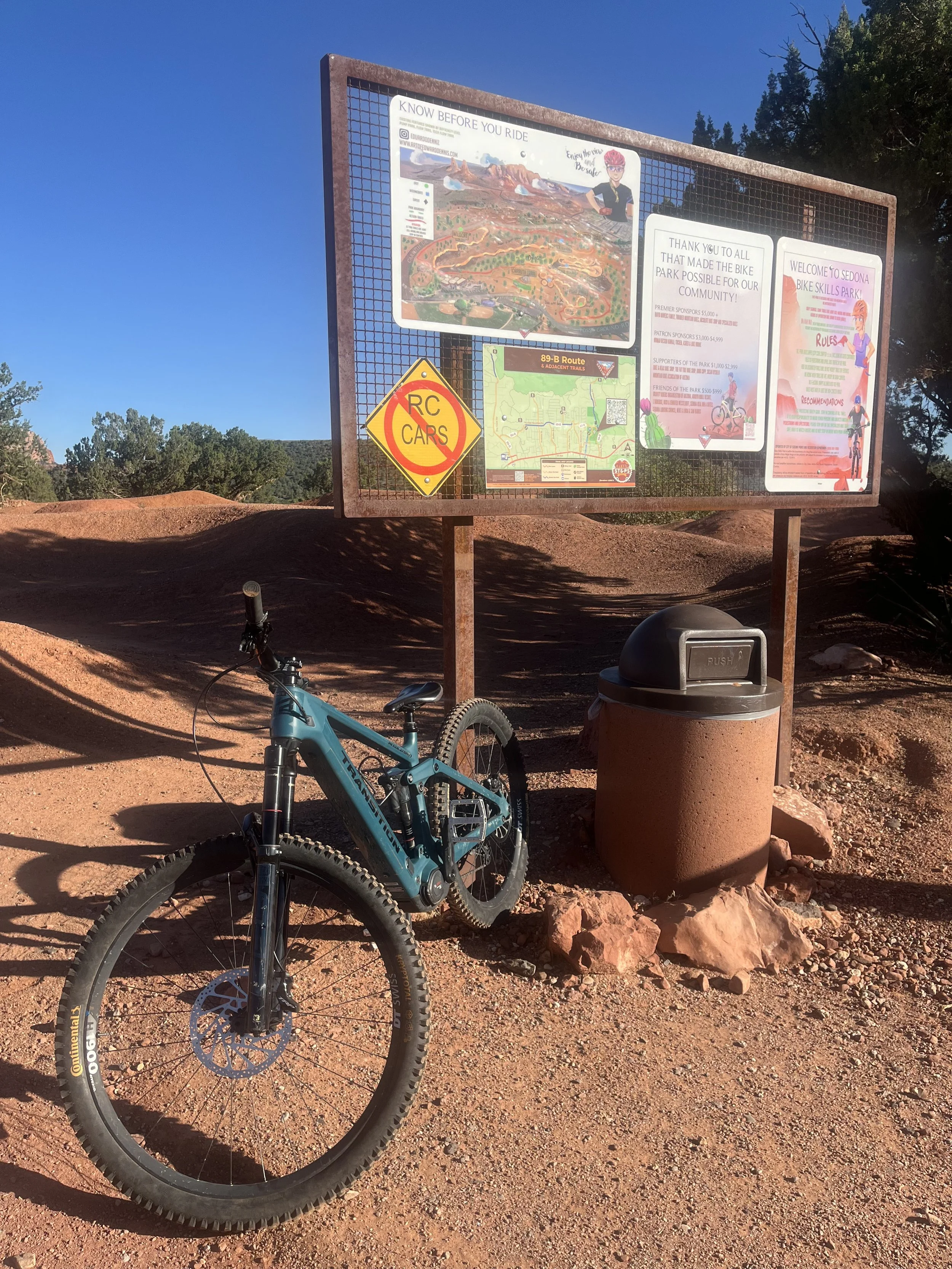 A blue mountain bike is parked on dirt ground in front of a large park information board. The board features trail maps, rules, and acknowledgments. A trash can is nearby, and trees and blue sky are visible in the background.