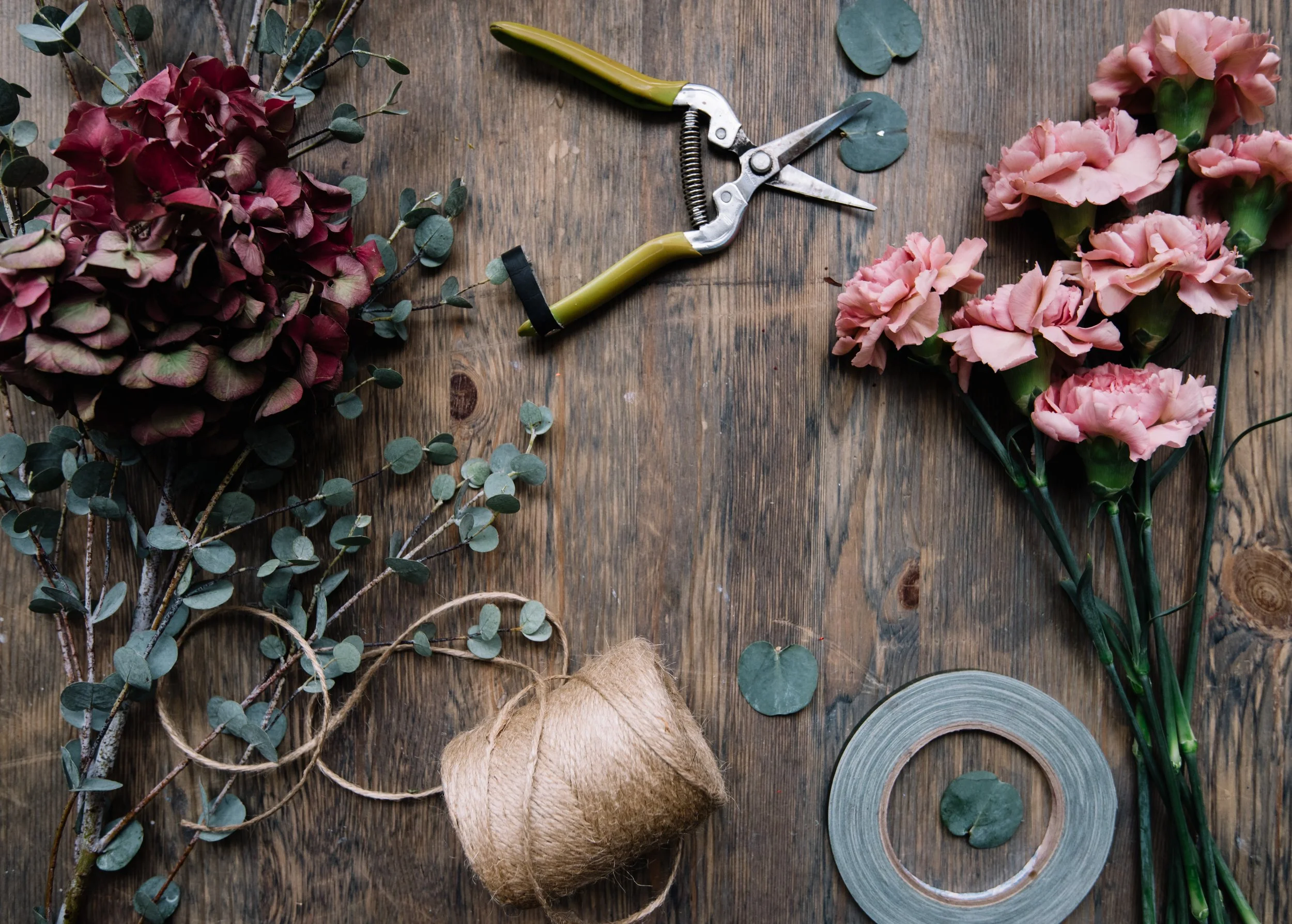 Various flowers and gardening tools on a wooden surface, including pink carnations, dried eucalyptus leaves, a ball of twine, a roll of gardening tape, and pruning shears.