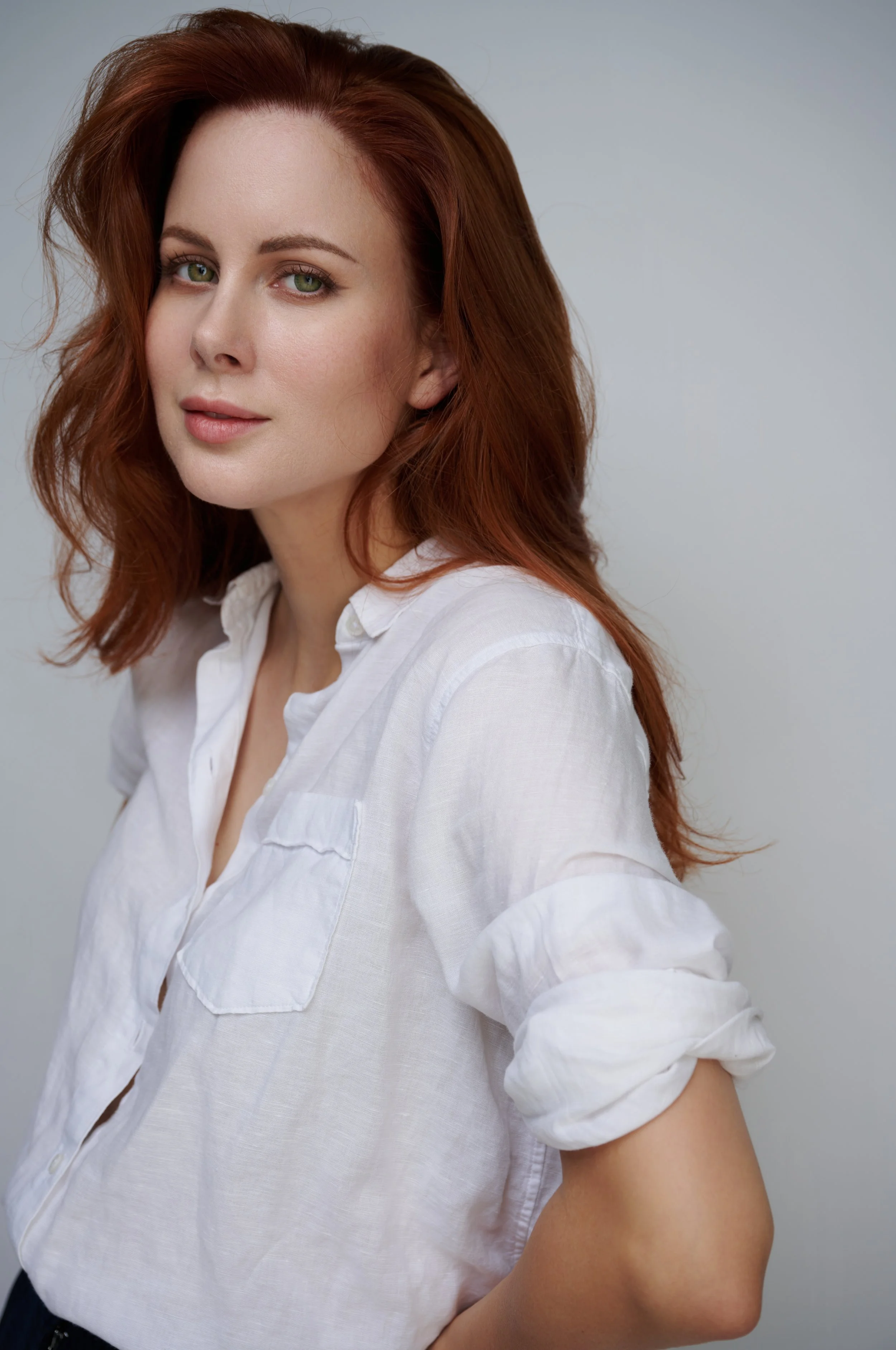 Close-up of a woman with red hair and green eyes wearing a white shirt against a plain background.