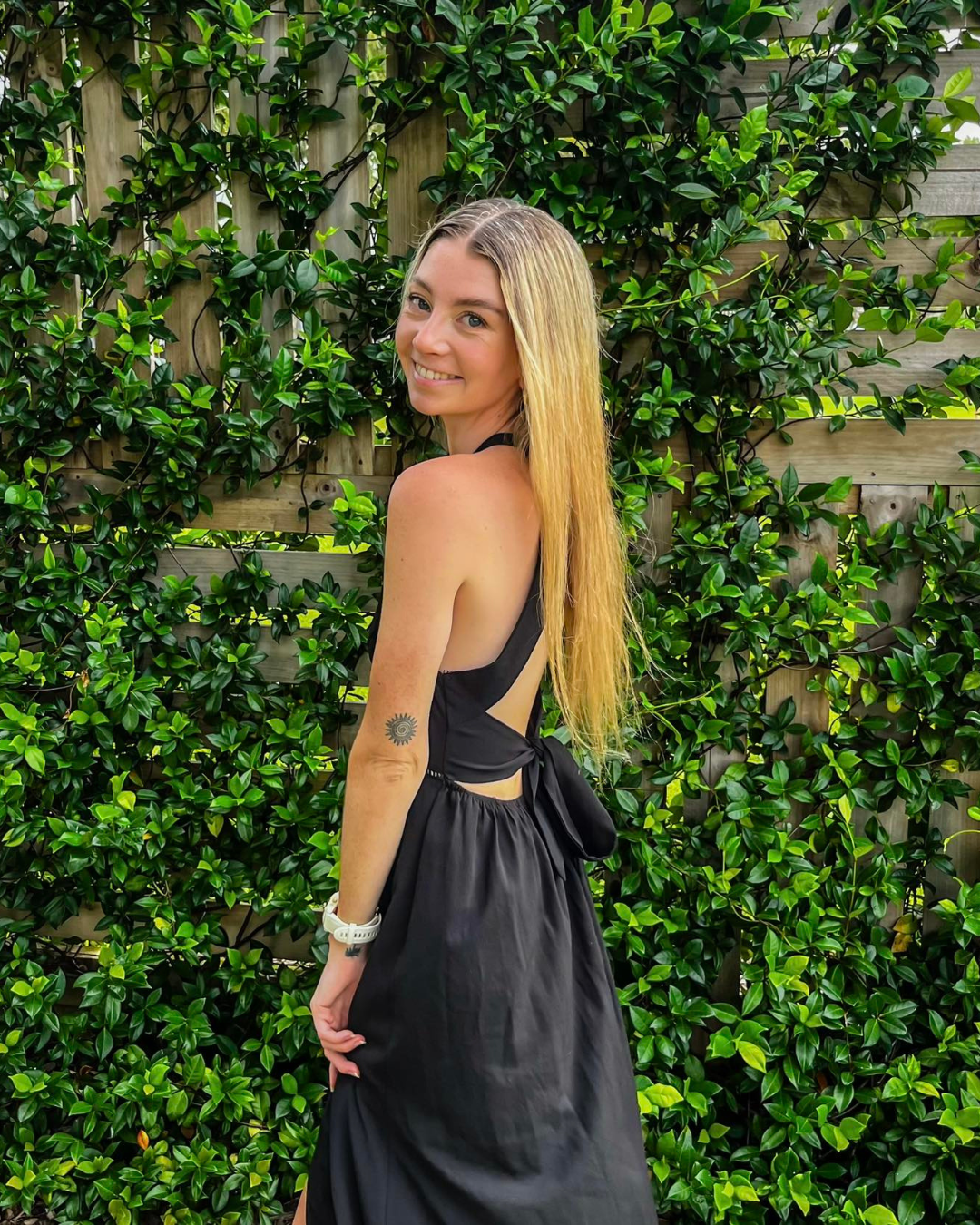 A young woman with long blonde hair smiling, wearing a black sleeveless dress with an open back, standing outdoors in front of a green leafy hedge and wooden fence.