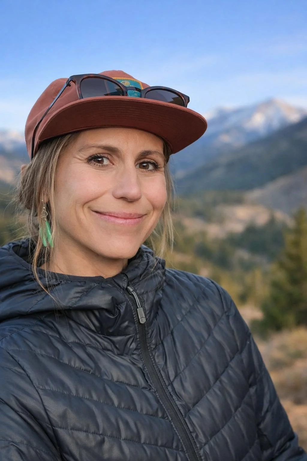 A woman wearing a brown cap with sunglasses on top, a dark puffy jacket, and turquoise earrings, smiling outdoors with mountains and trees in the background.