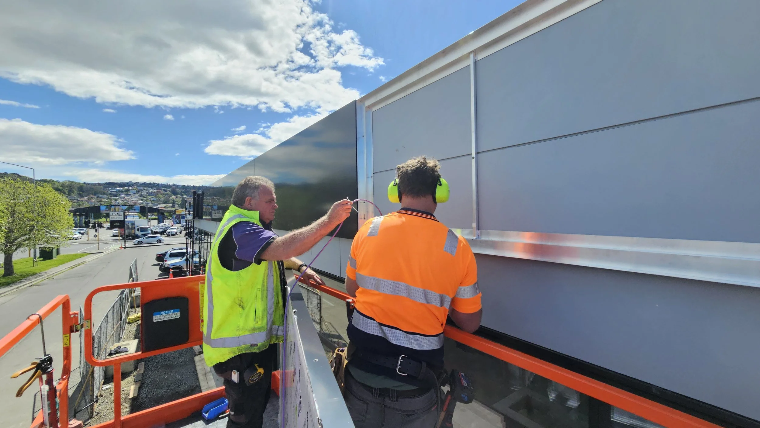 Two signage installers on a lift working on a metal wall panel outside on a bright, partly cloudy day.