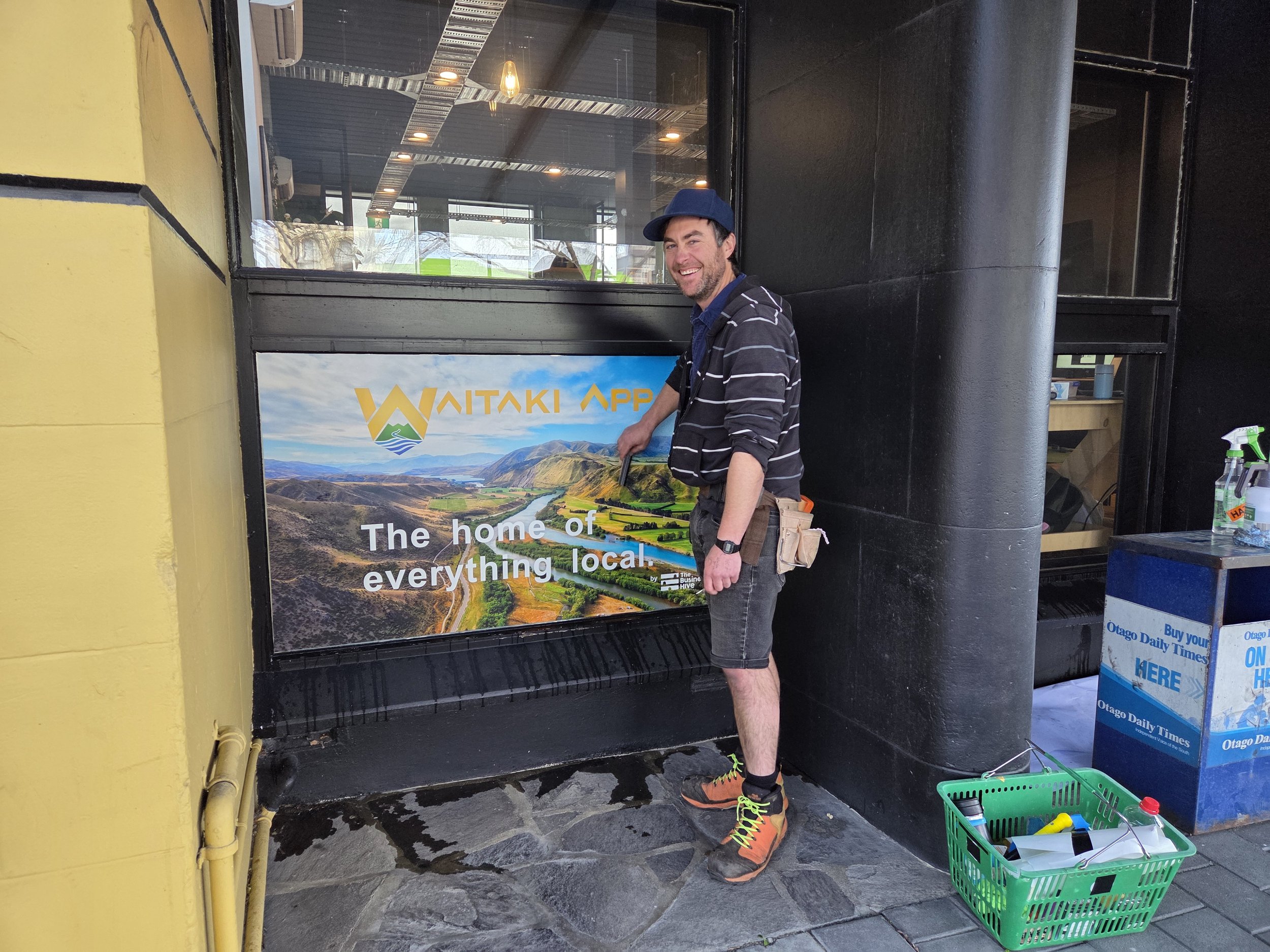 A man in outdoor gear installs a window graphic hat reads 'Waitaki App, The home of everything local,' standing outside a building with a large window and black exterior walls.