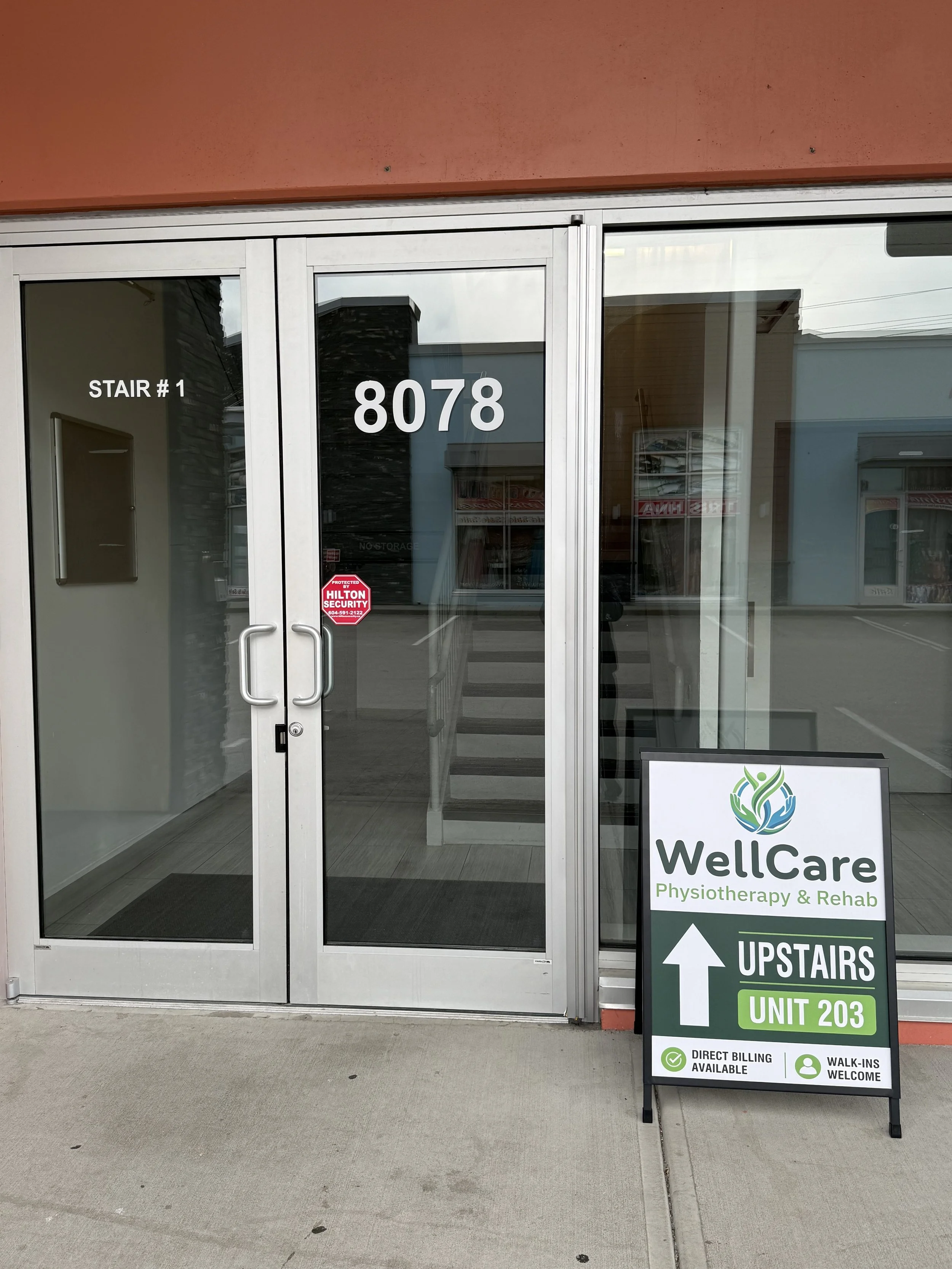 Store entrance with glass doors, signage for WellCare Physiotherapy and Rehab, a black and white standing sign indicating upstairs, unit 203, and services like direct billing and walk-ins welcome.