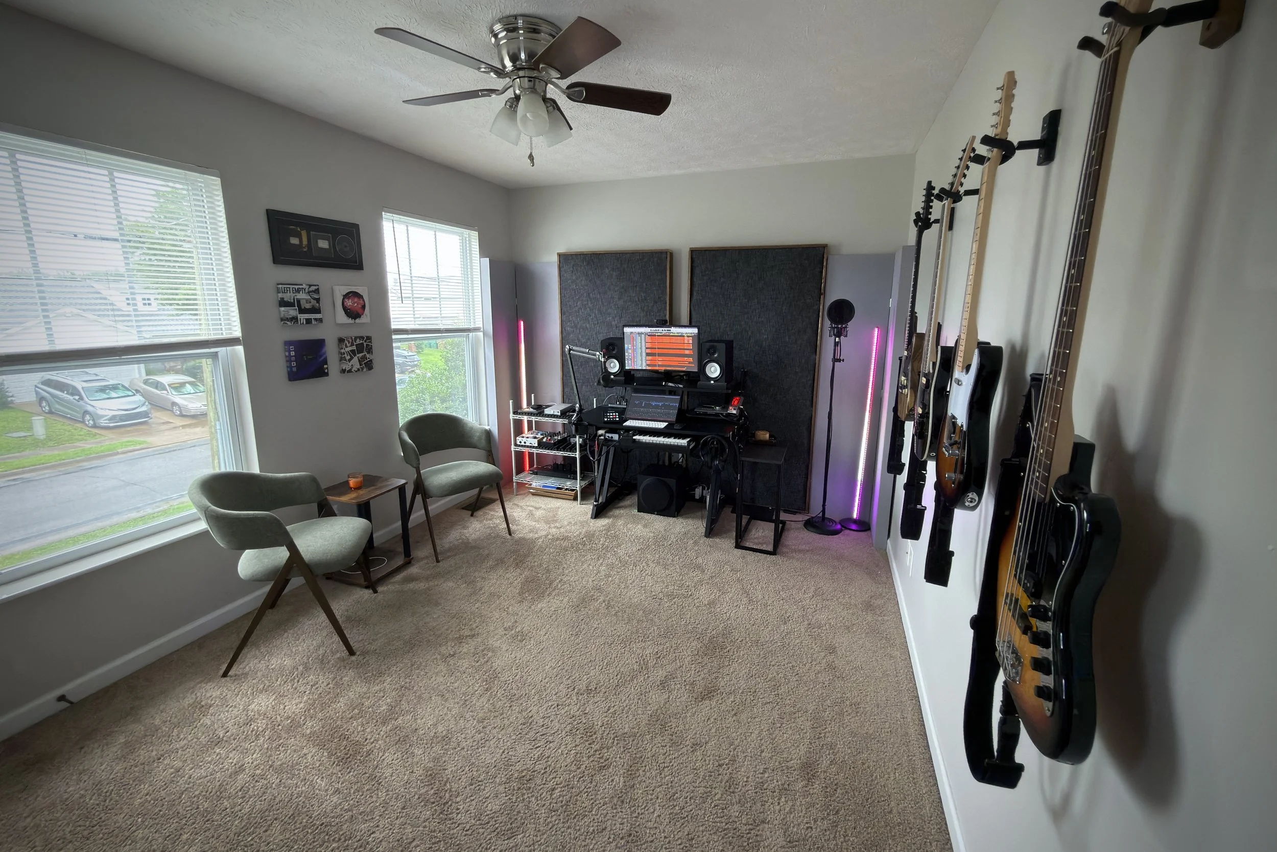 A room with multiple guitars hanging on the wall, a music production setup with monitors, keyboard, microphone, and LED lights, two chairs, a window with blinds, and vinyl record wall art.