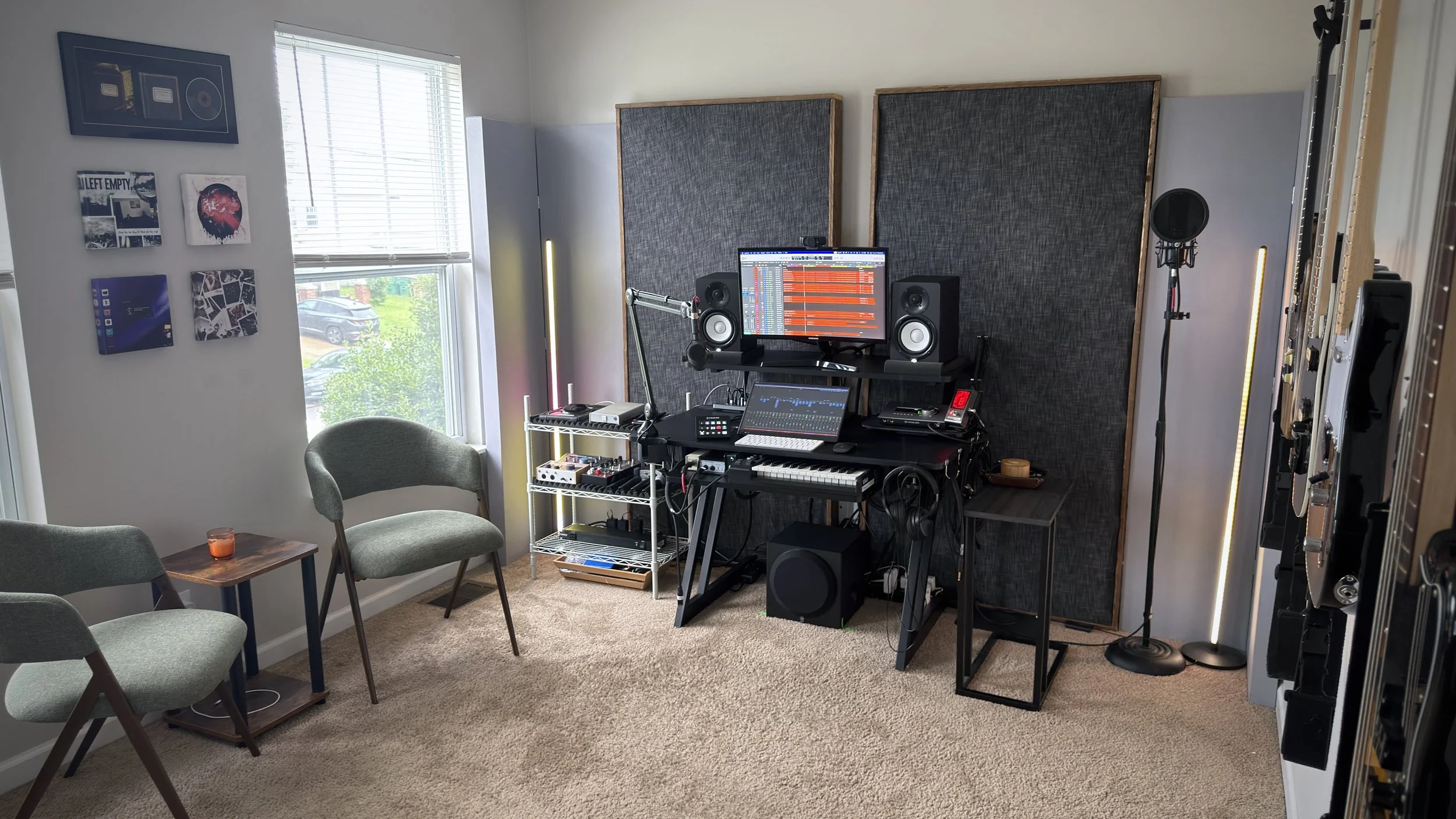 A music recording studio with a computer monitor displaying audio editing software, two studio speakers, a microphone on a stand, and soundproof panels on the wall. There are two chairs and a small table with a candle, and a window with blinds showing a parking lot outside.