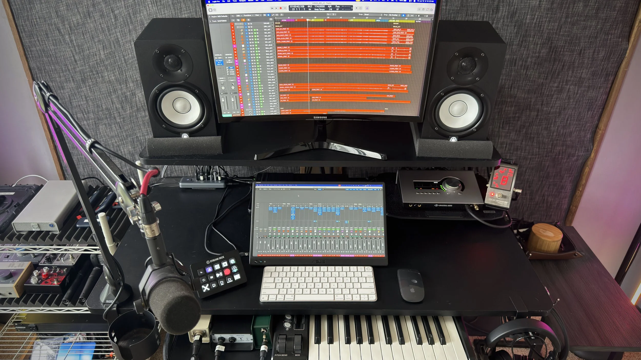 A music production workstation featuring a computer monitor displaying a digital audio workstation with multiple tracks, studio speakers on either side, a MIDI keyboard, a tablet showing a sound mixer, a microphone, and various audio equipment, set against a textured wall background.