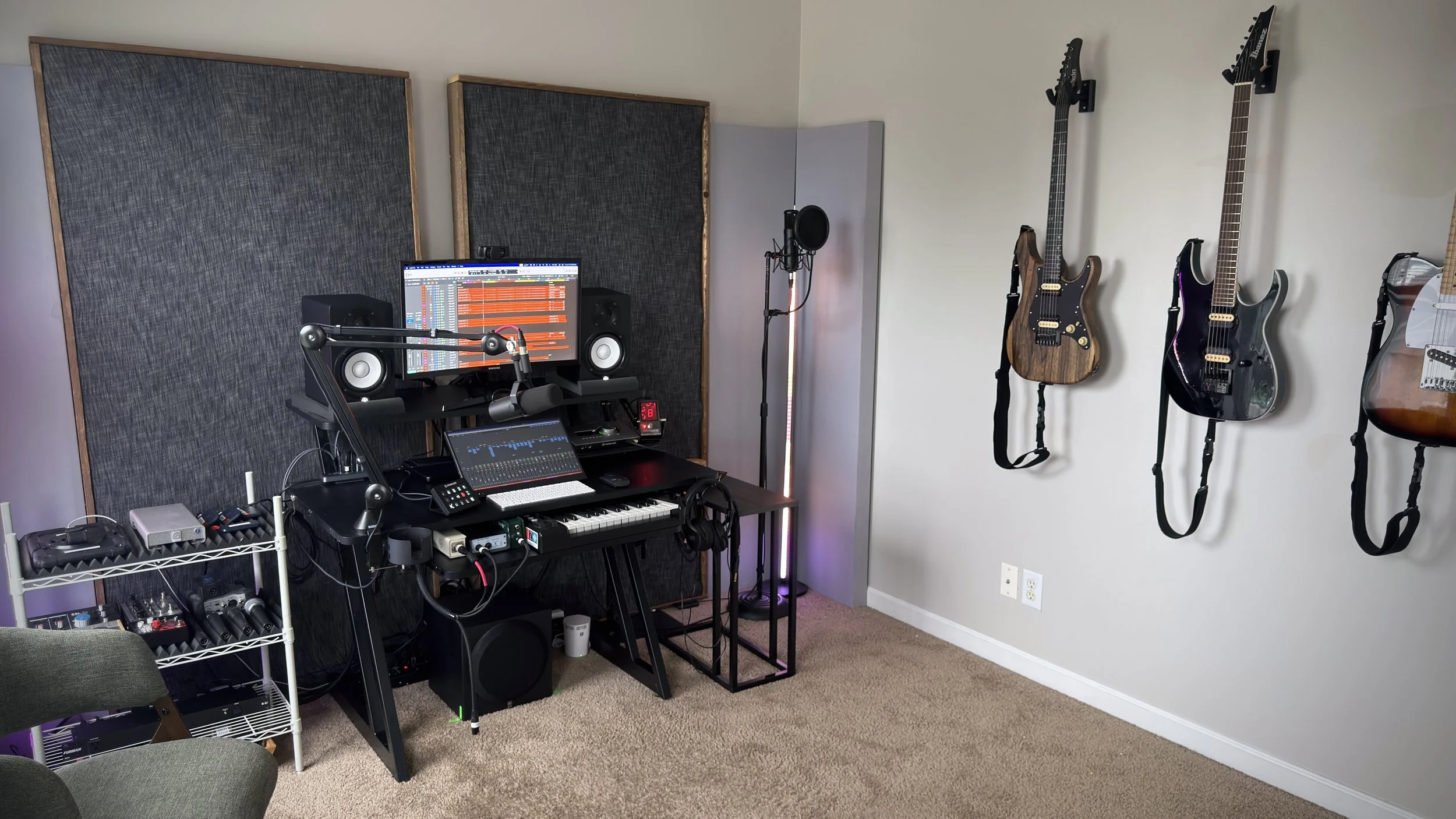 Home recording studio with a desk holding a computer monitor, studio speakers, MIDI keyboard, microphone on a stand, and audio equipment. Several electric guitars are mounted on the wall.