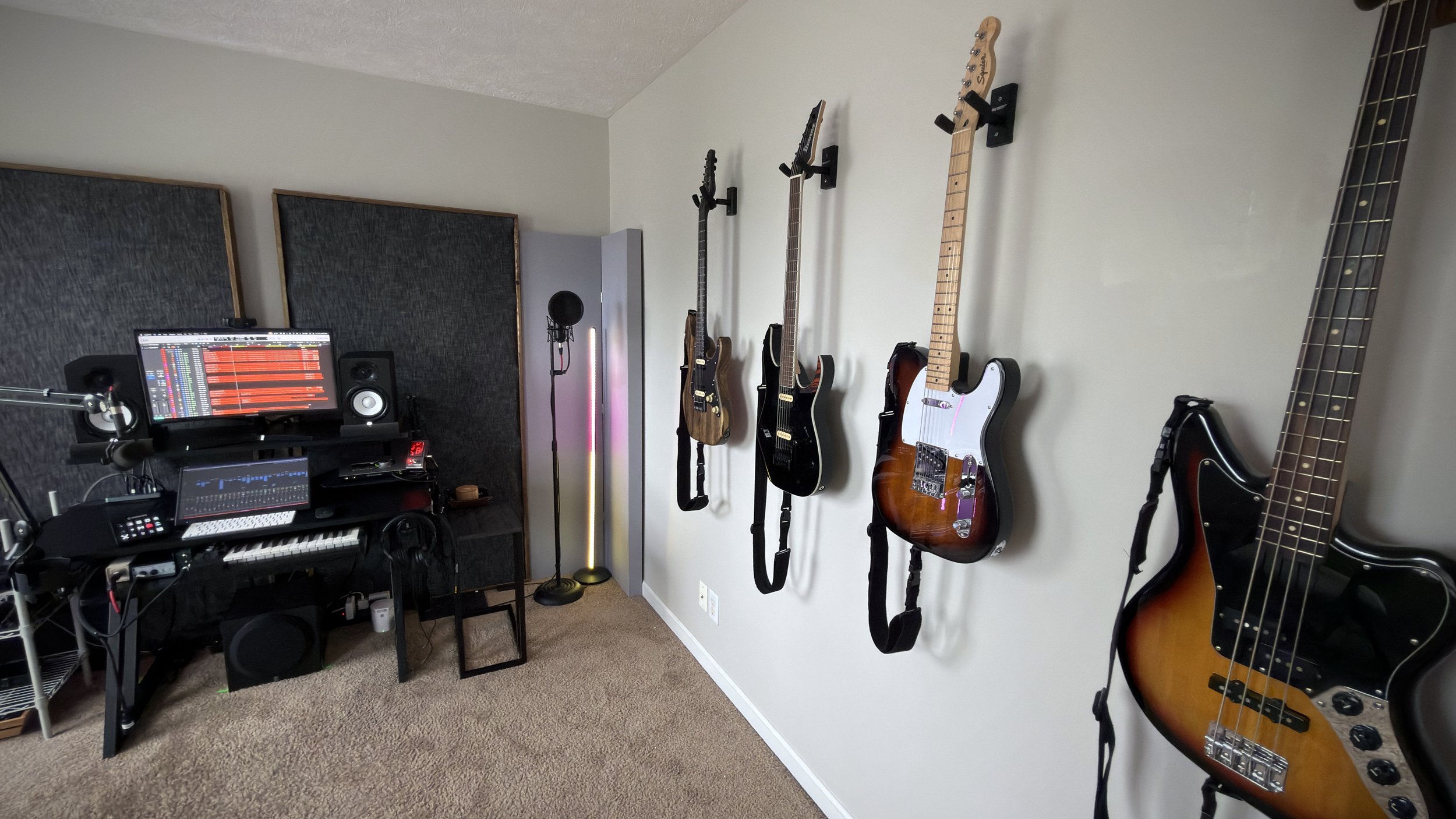 A music studio room with four guitars hanging on the wall, a computer with audio editing software on a desk, and studio soundproofing panels.