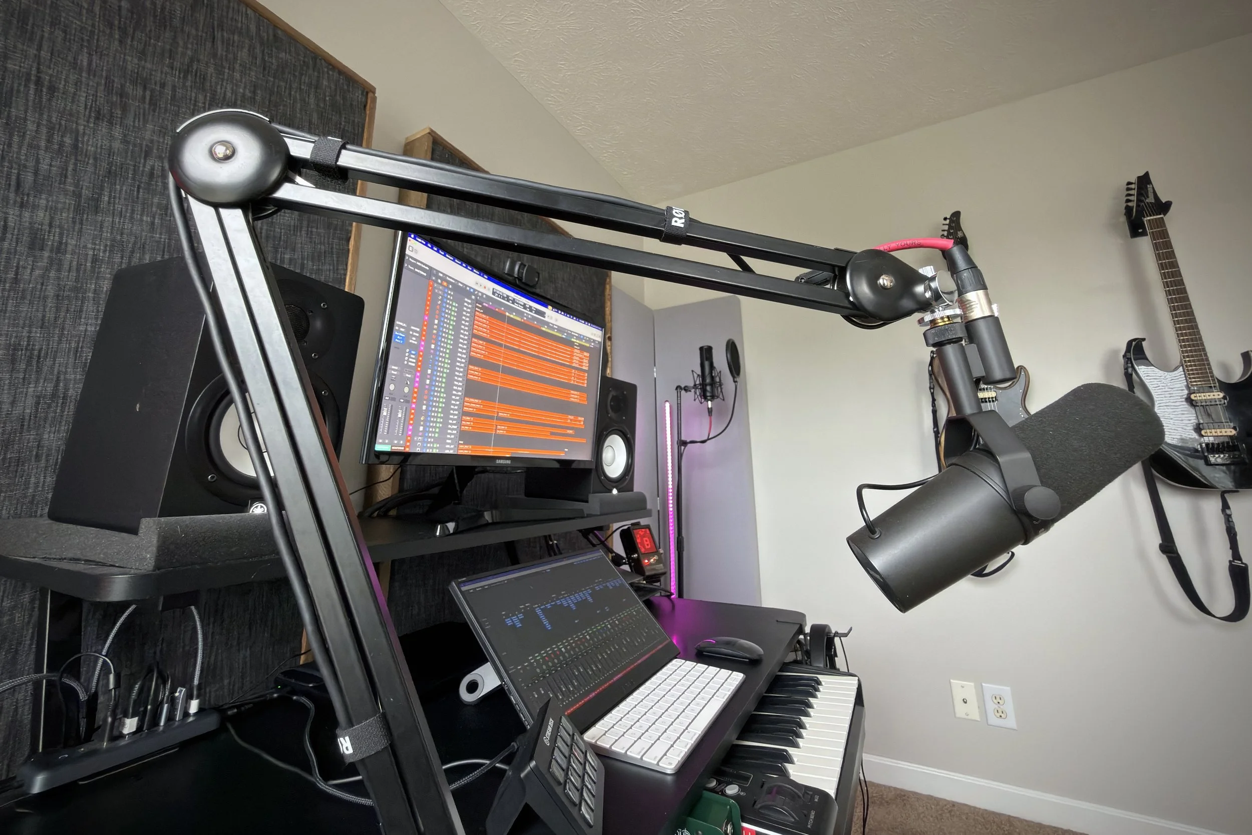 A home music studio setup with a computer monitor displaying digital audio workstation software, a keyboard, studio monitor speakers, a microphone with a pop filter, an acoustic guitar hanging on the wall, and various audio equipment on a desk and shelves.