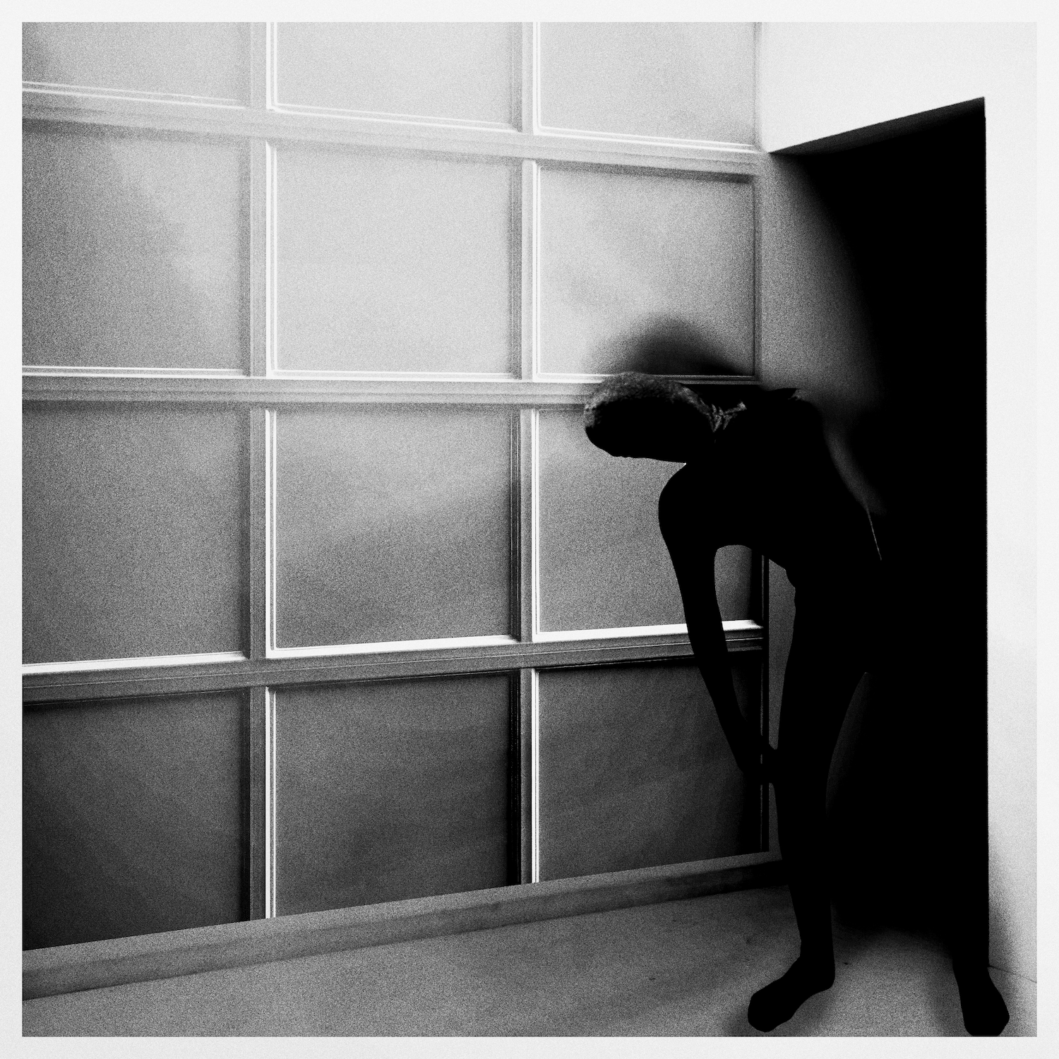 Black and white photo of a person with short hair, bending over in front of a large shelf or cabinet with glass doors, illuminated by a soft light.
