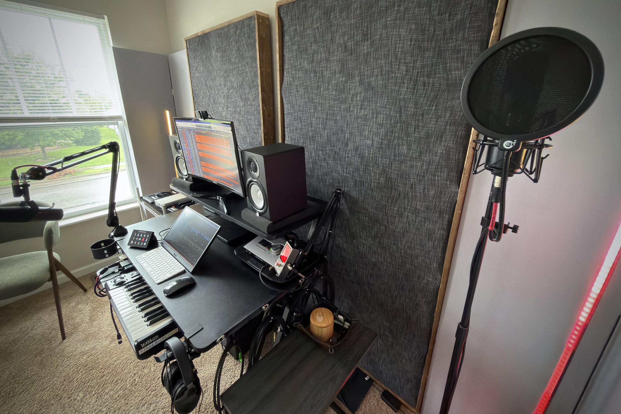 A music studio room with a computer monitor, studio speakers, a keyboard, and a microphone on a stand, with soundproofing panels on the wall and a large window showing greenery outside.